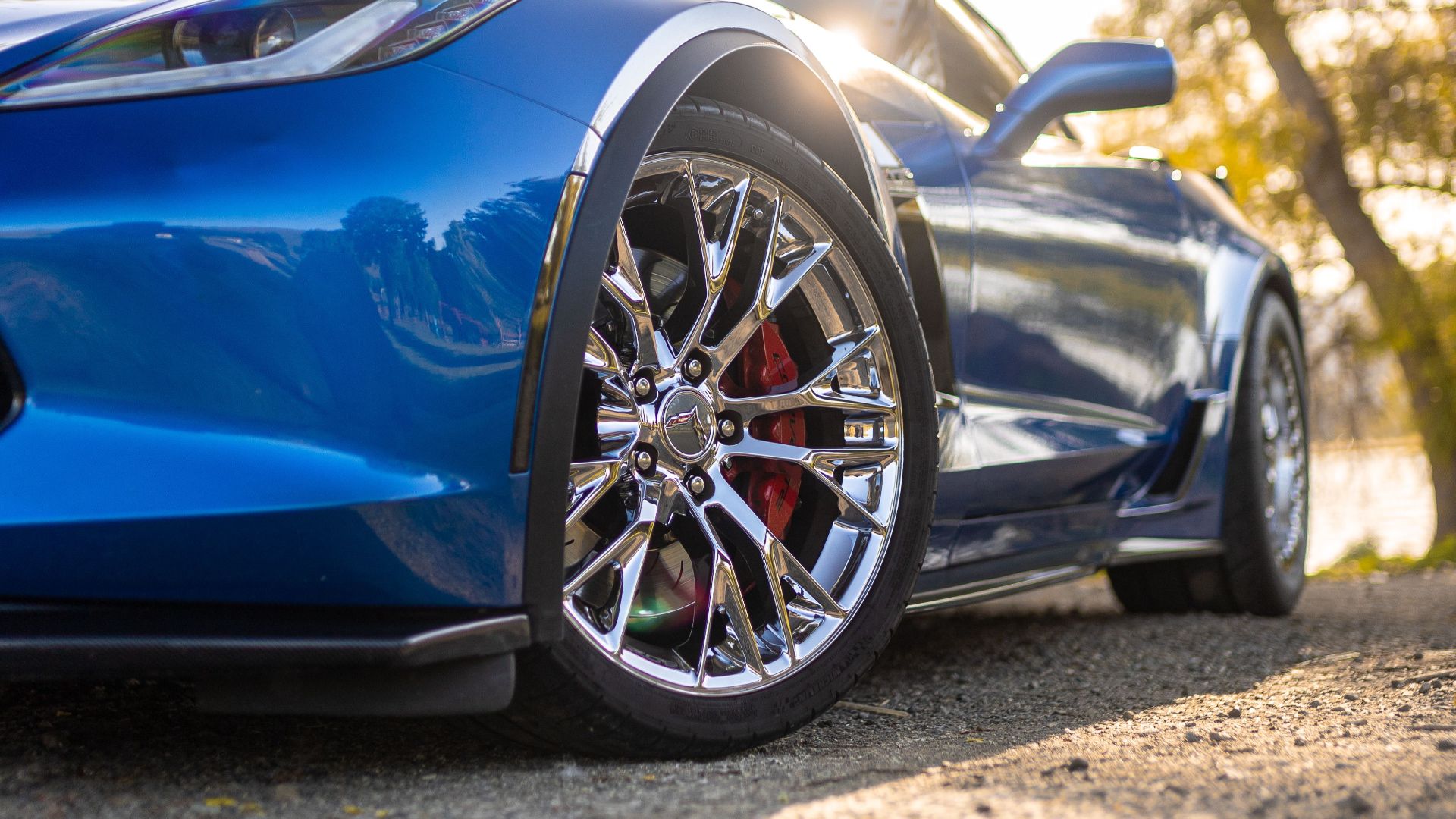 Vibrant blue Chevrolet Corvette sports car with shiny alloy wheels parked in a scenic rural setting with sunlight filtering through trees.