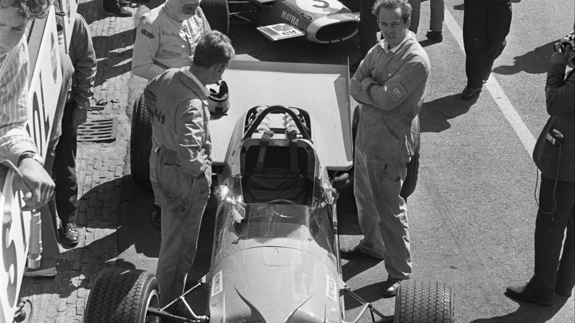 Bruce McLaren (centre left, white balaclava) prepares to take his seat in his McLaren M7C Formula One car, prior to the 1969 Dutch Grand Prix. Behind, French driver Jean-Pierre Beltoise (helmet on) is about to board his Matra MS80.