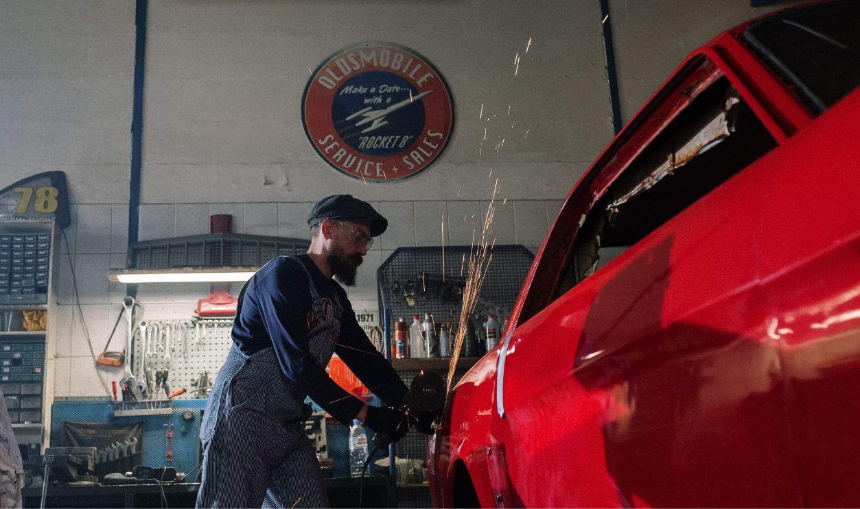 A bearded auto mechanic using a grinder on a red car in a vintage garage