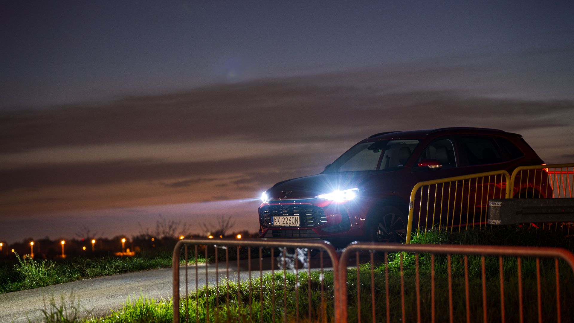 A red SUV with headlights on parked along a rural road at sunset in Kokotów, Poland.