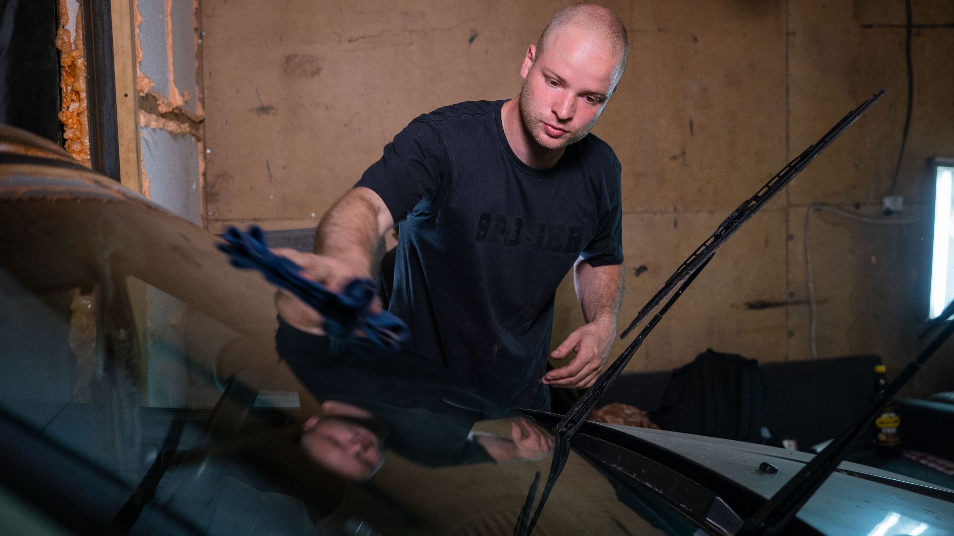 A man wipes a car windshield inside a garage, focused on cleaning.