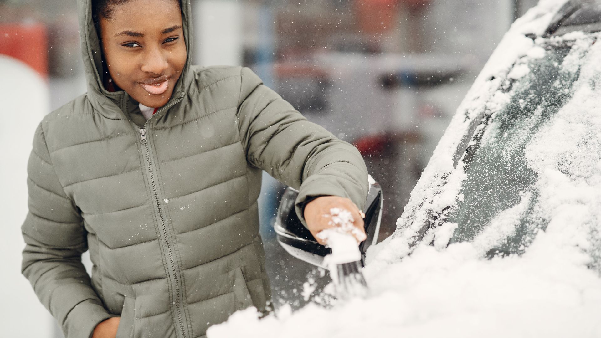 A young woman wearing a jacket clears snow from her car during a winter day.