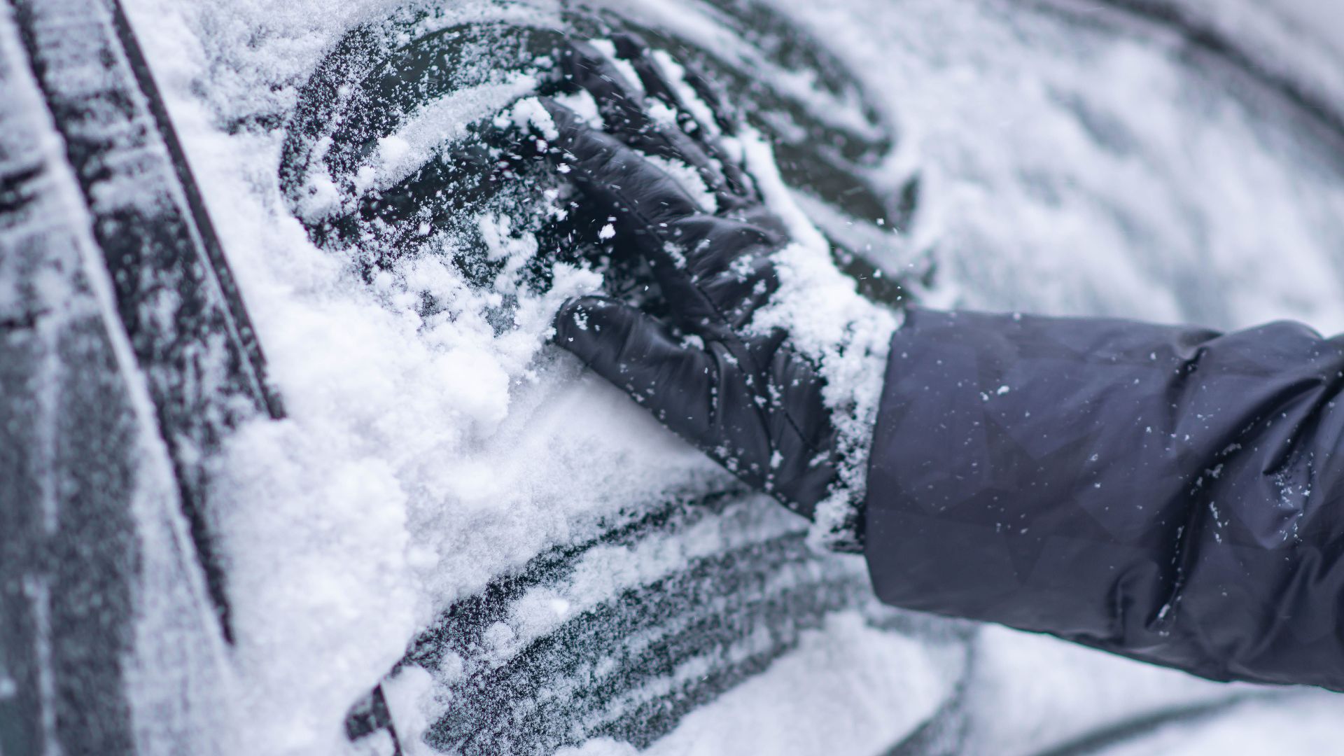 A close-up of a gloved hand clearing snow from a car window during winter.