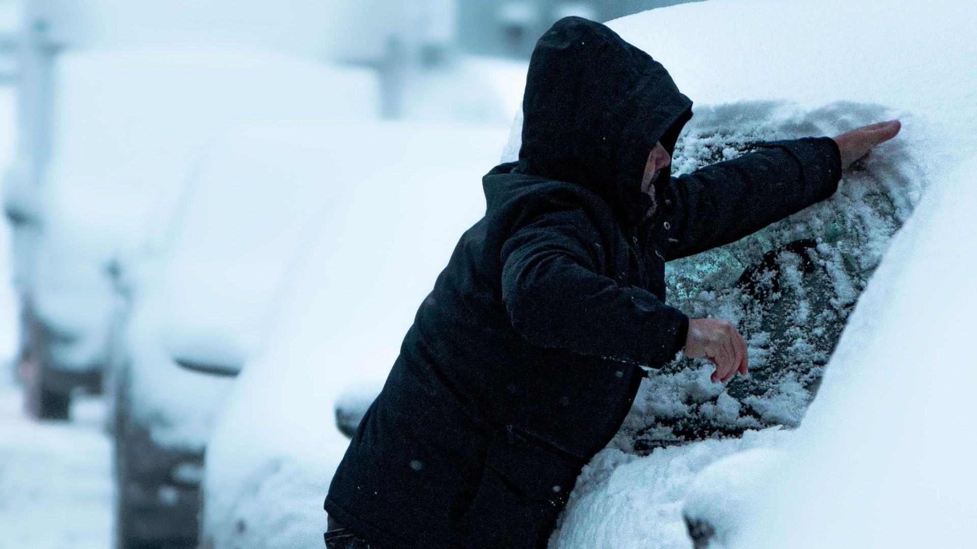 A man clears snow from a car parked on a wintery street, highlighting seasonal car maintenance.