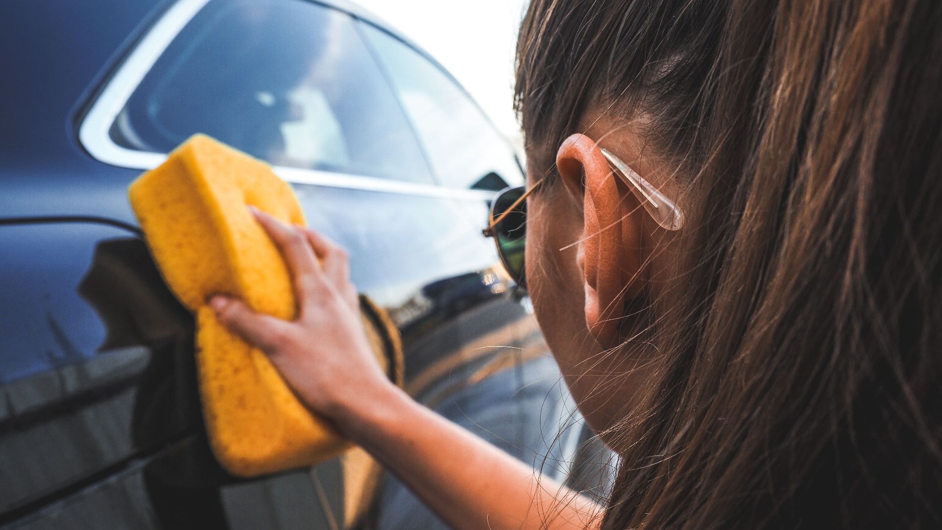 Close-up of a woman handwashing a black car using a yellow sponge, highlighting cleaning process.