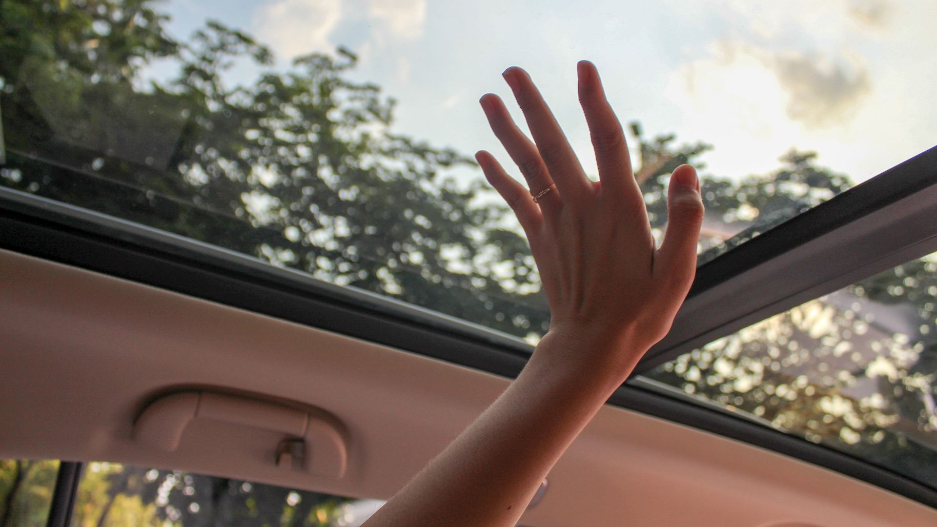 A hand reaching out through a car sunroof, touching the sky on a sunny day.