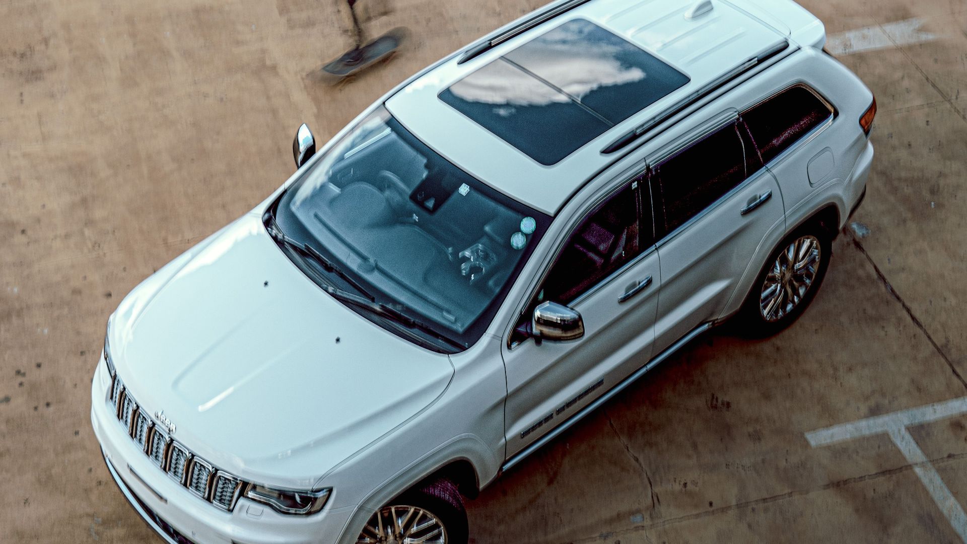 Aerial view of white SUV in a sparse parking area with a skateboarder in motion nearby.