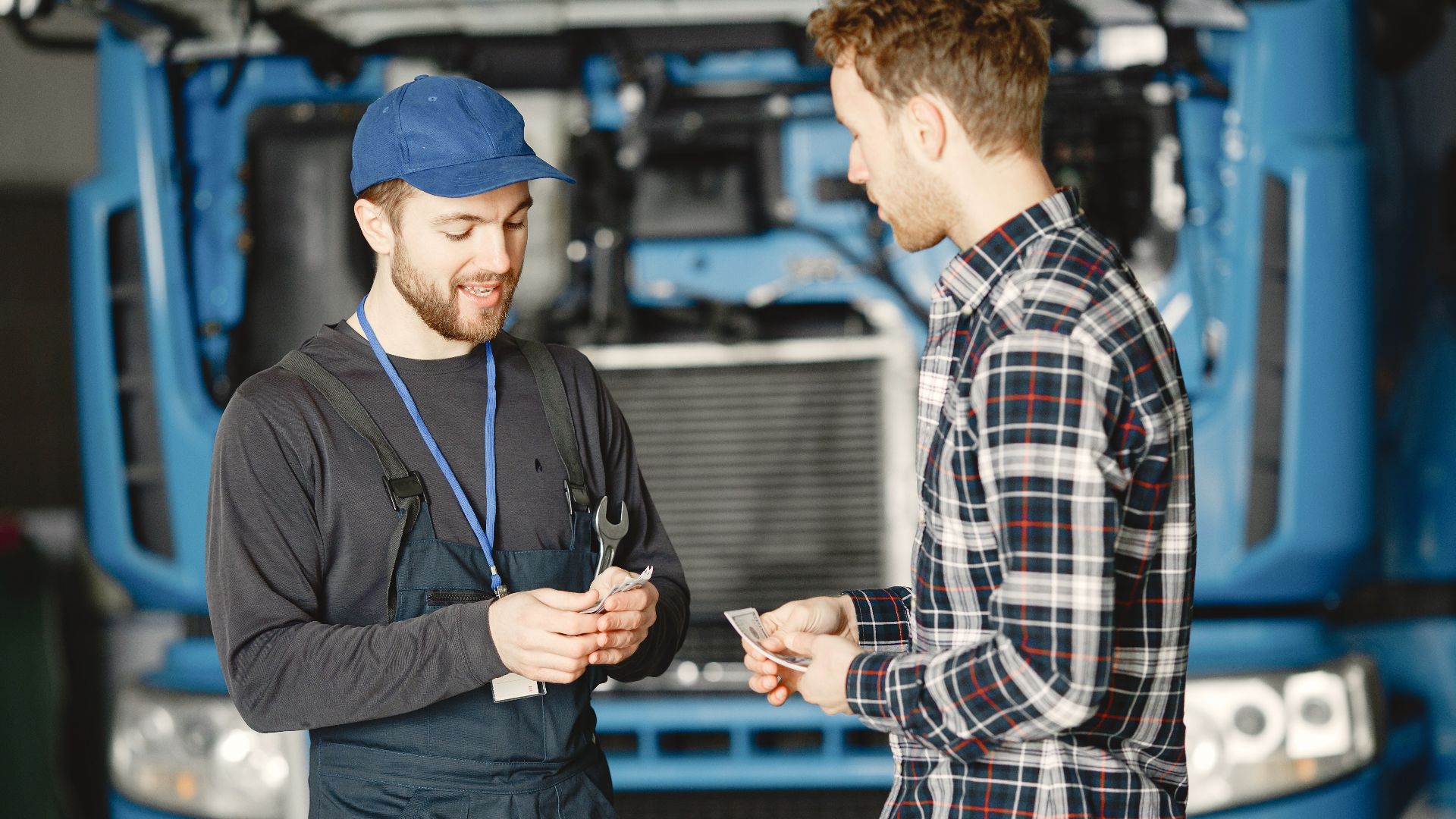 Auto mechanic in blue uniform receives payment from customer in a garage setting with a truck in the background.