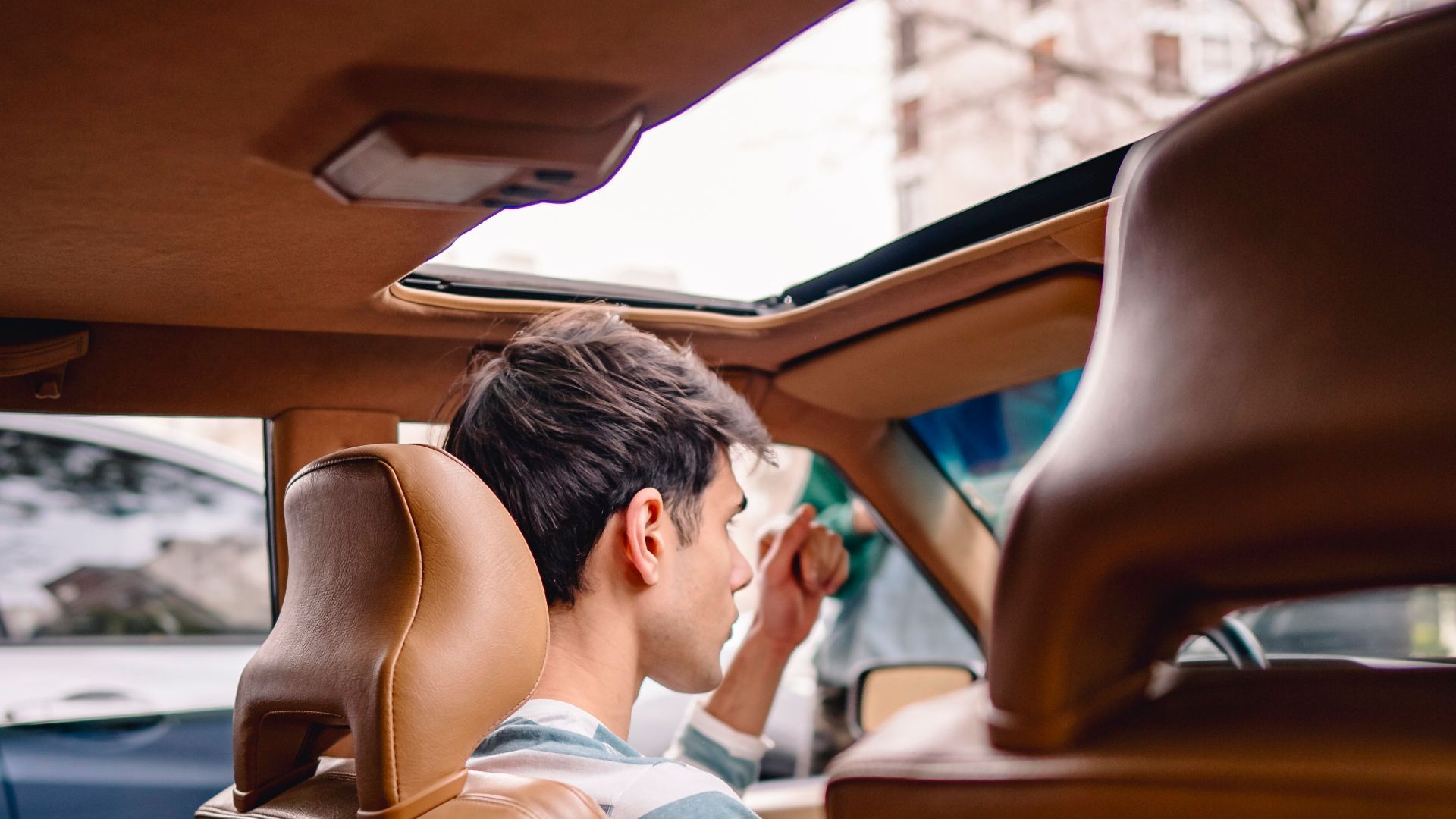 Back view of a young man sitting in a vintage car with a sunroof, enjoying a sunny day.