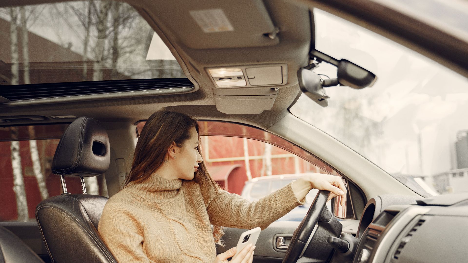 From window side view of adult woman in warm clothes sitting at wheel of modern car waiting for friend and using smartphone