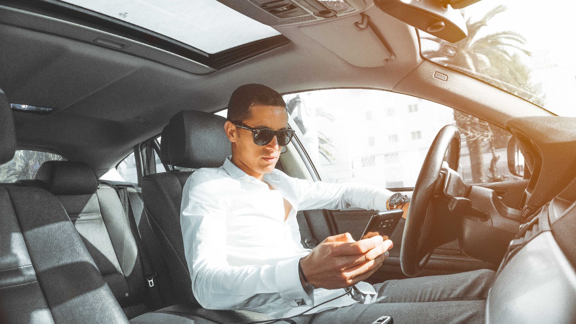 A man wearing sunglasses texts on his phone while driving a car in Morocco.