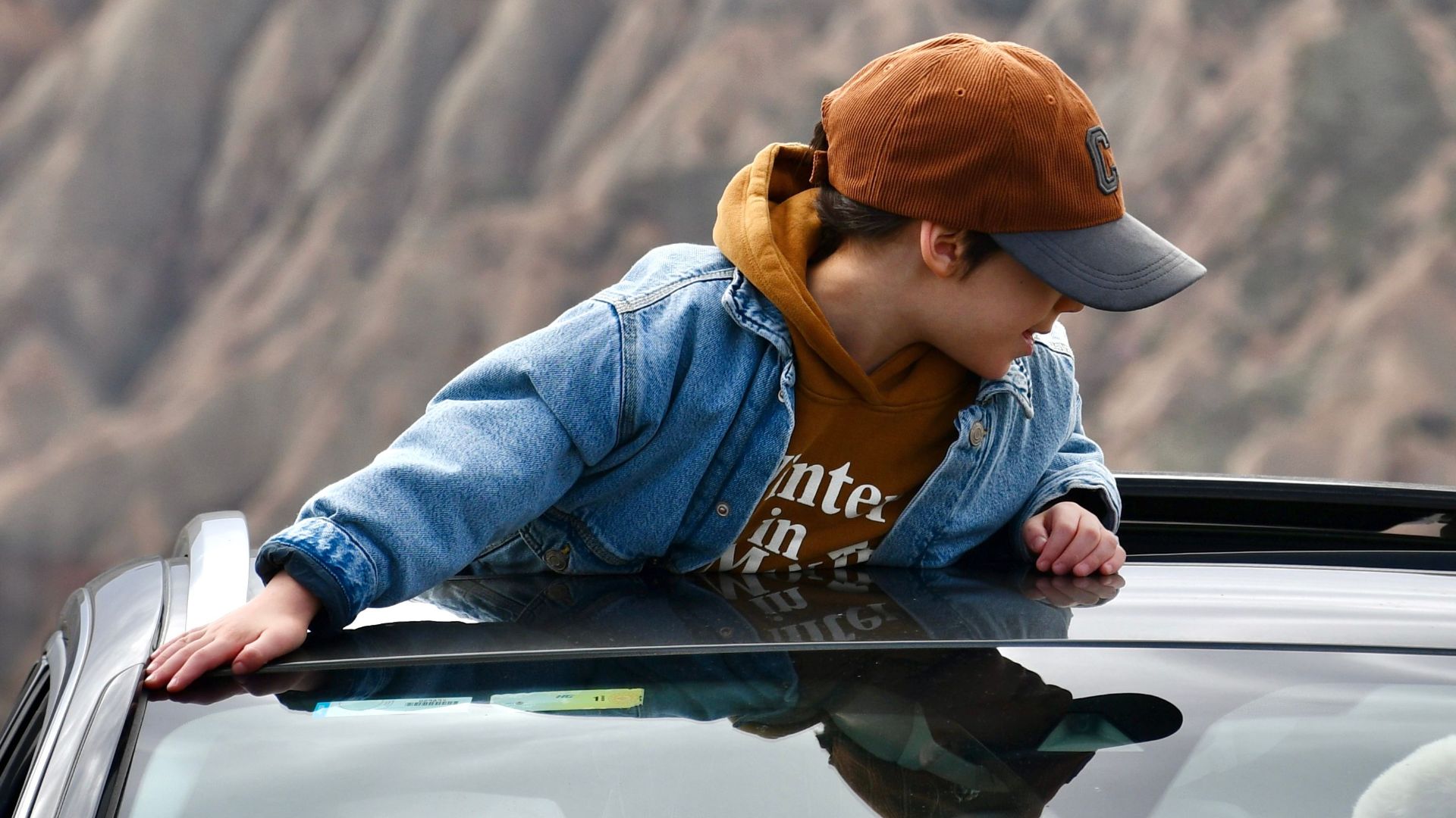 A young person in denim leans out of a car sunroof, taking in the scenic mountain view.