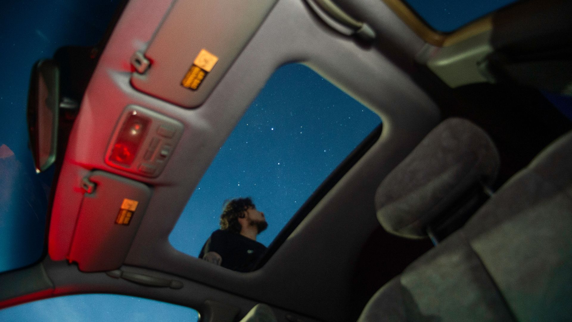 Man stargazing through car sunroof, capturing a serene night sky with stars.