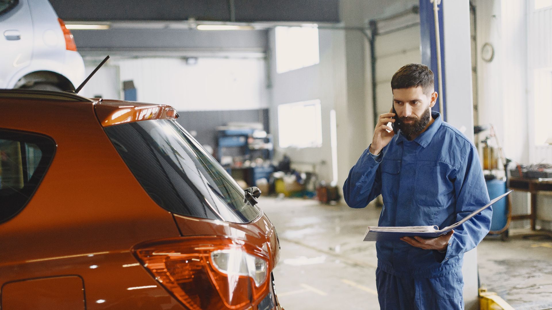 Mechanic in a blue uniform talks on phone beside a red car in an auto repair shop.