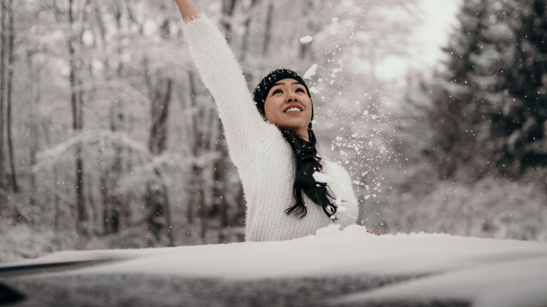 Cheerful ethnic female throwing snow above head near auto against trees in forest during snowfall