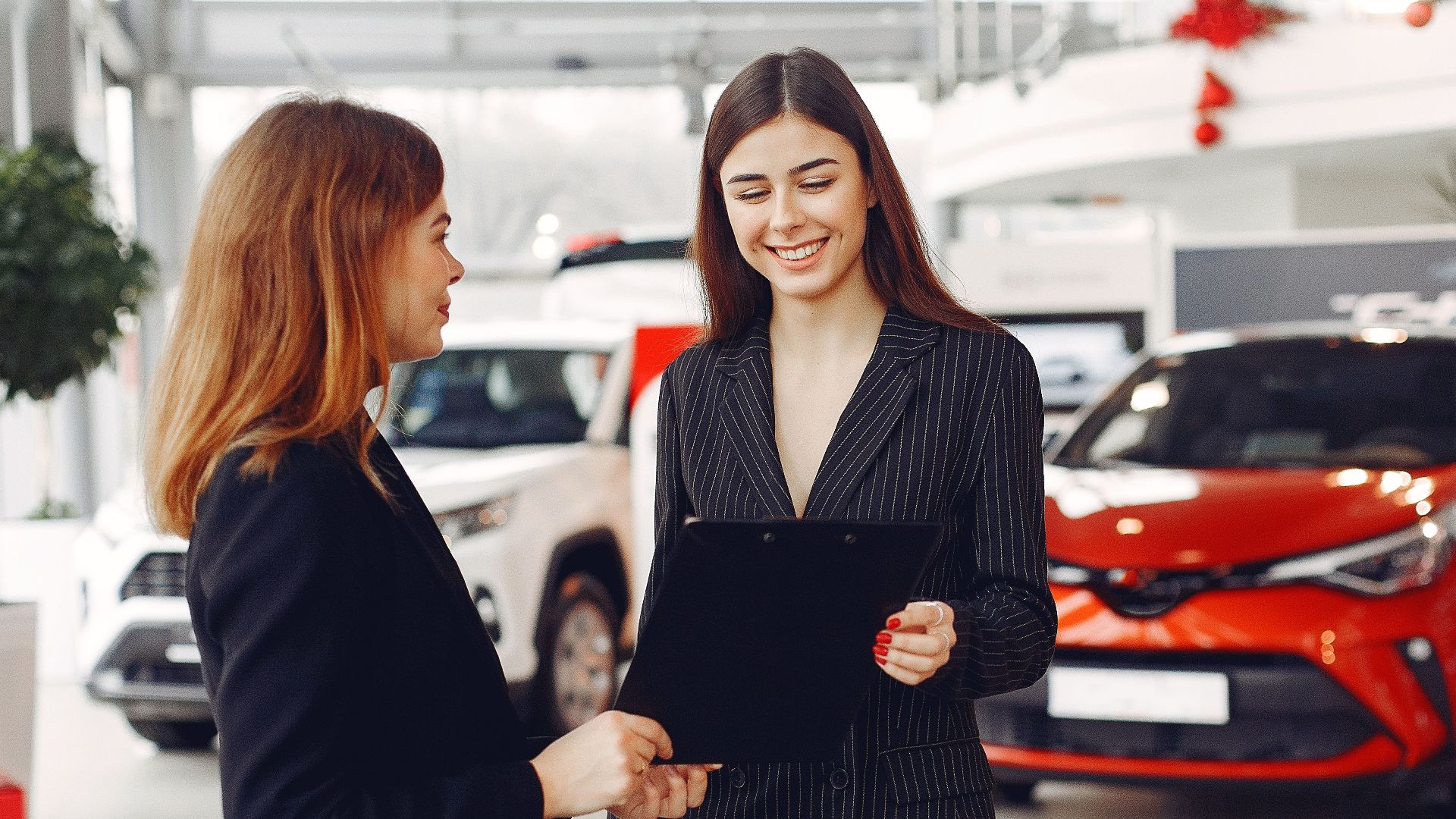Cheerful young friendly dealer in formal stylish black dress showing contract to smiling female customer in black jacket while standing in car showroom against new shiny automobiles