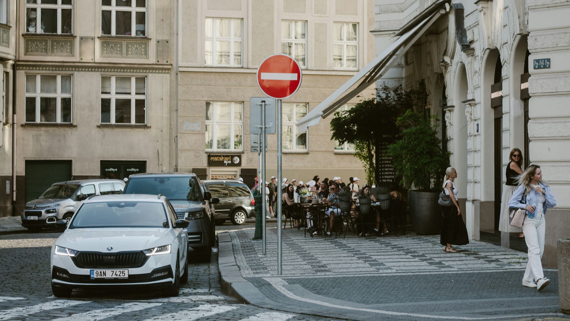 City street with cars, no entry sign, and outdoor cafe.