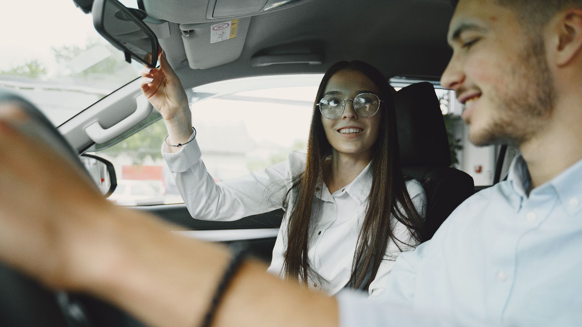 A cheerful young couple driving with focus on woman adjusting the mirror.