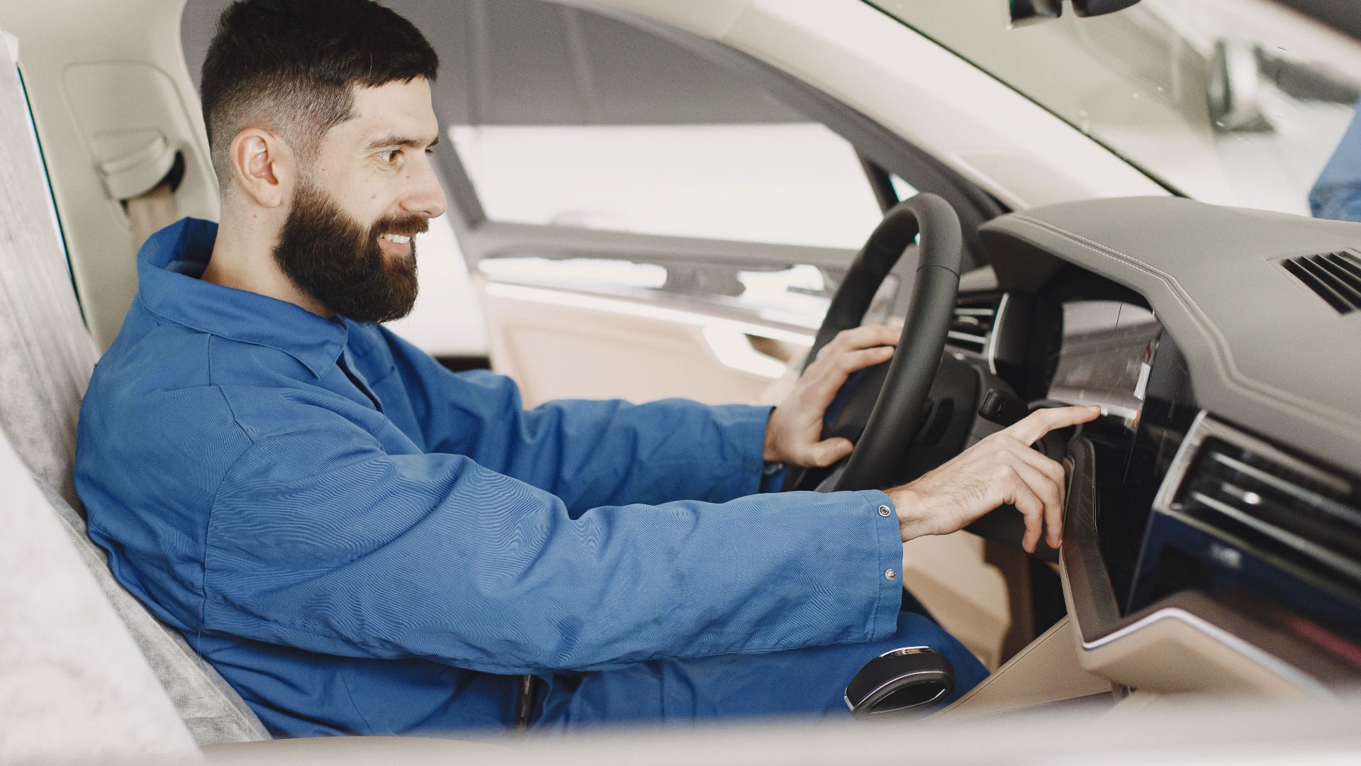 Mechanic in blue coveralls interacts with car dashboard, smiling and focused.