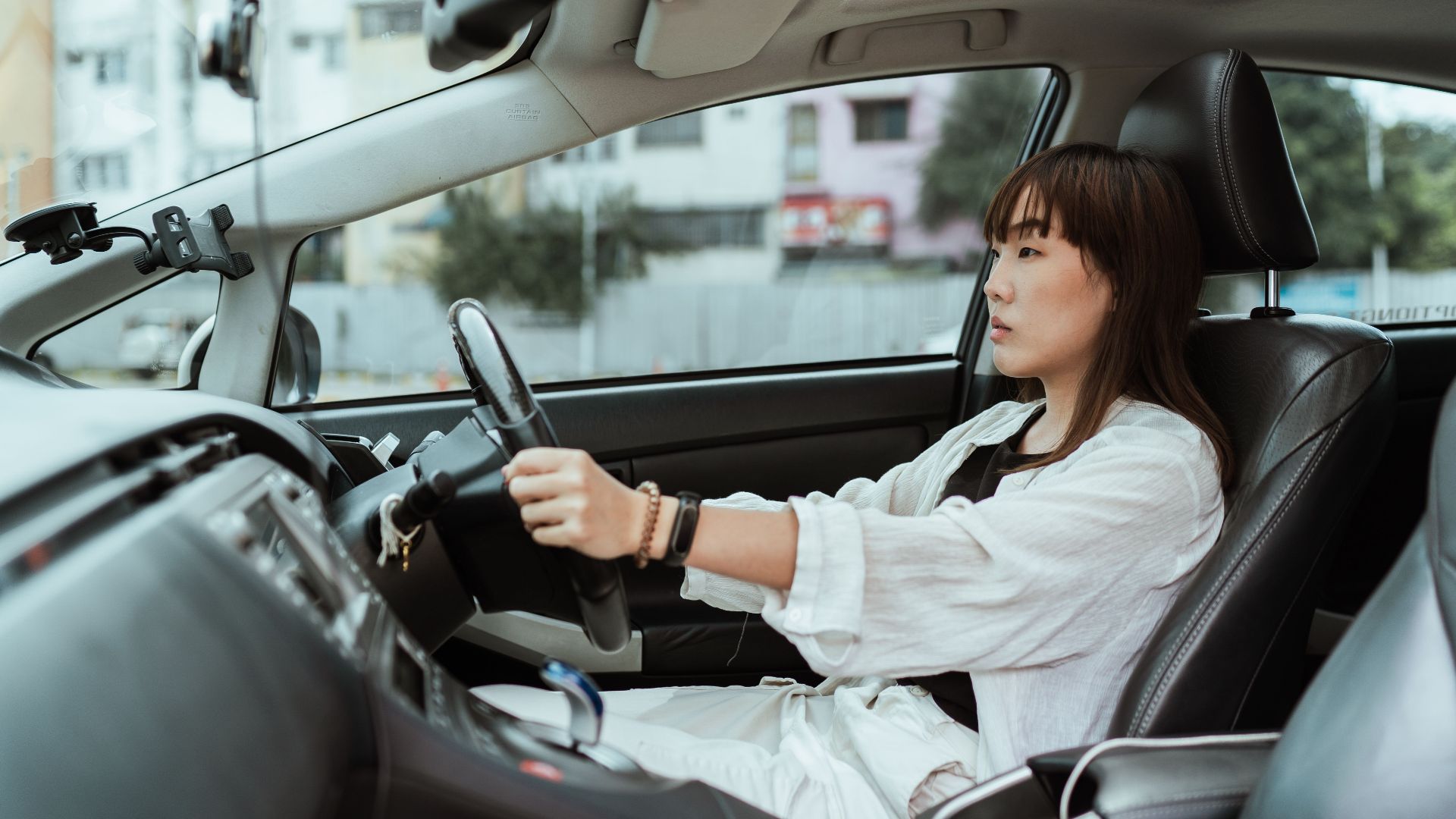 Side view unemotional Asian female in casual white clothes steering right hand drive car along urban streets in daytime