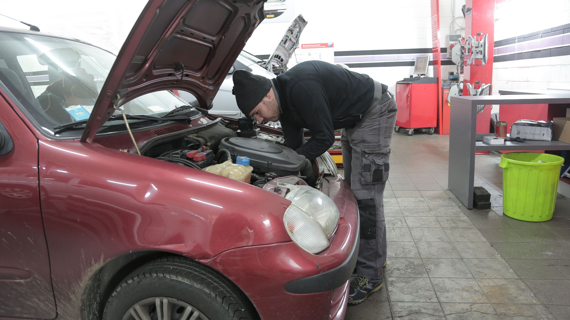 Mechanic performs engine inspection on a red car indoors, in a garage workshop.