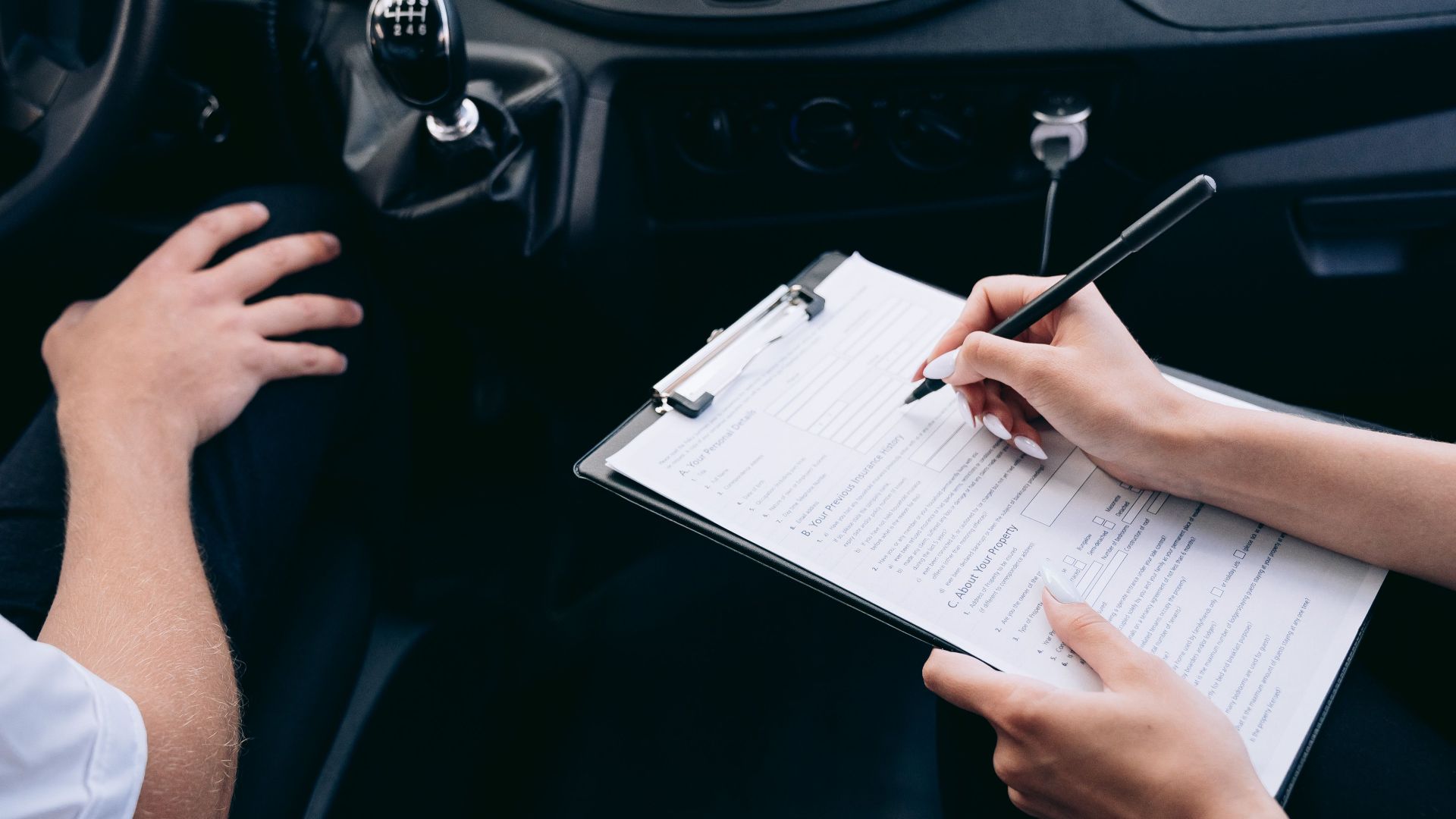 Close-up of a person writing on a clipboard inside a car, showing hands and a gear shift.