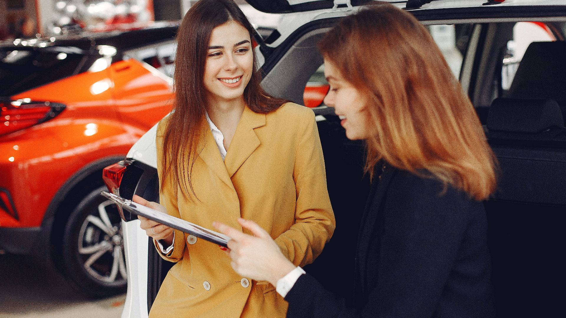 Positive smiling agent in trendy formal light brown suit discussing with happy excited female customer details of contract while leaning on car in showroom