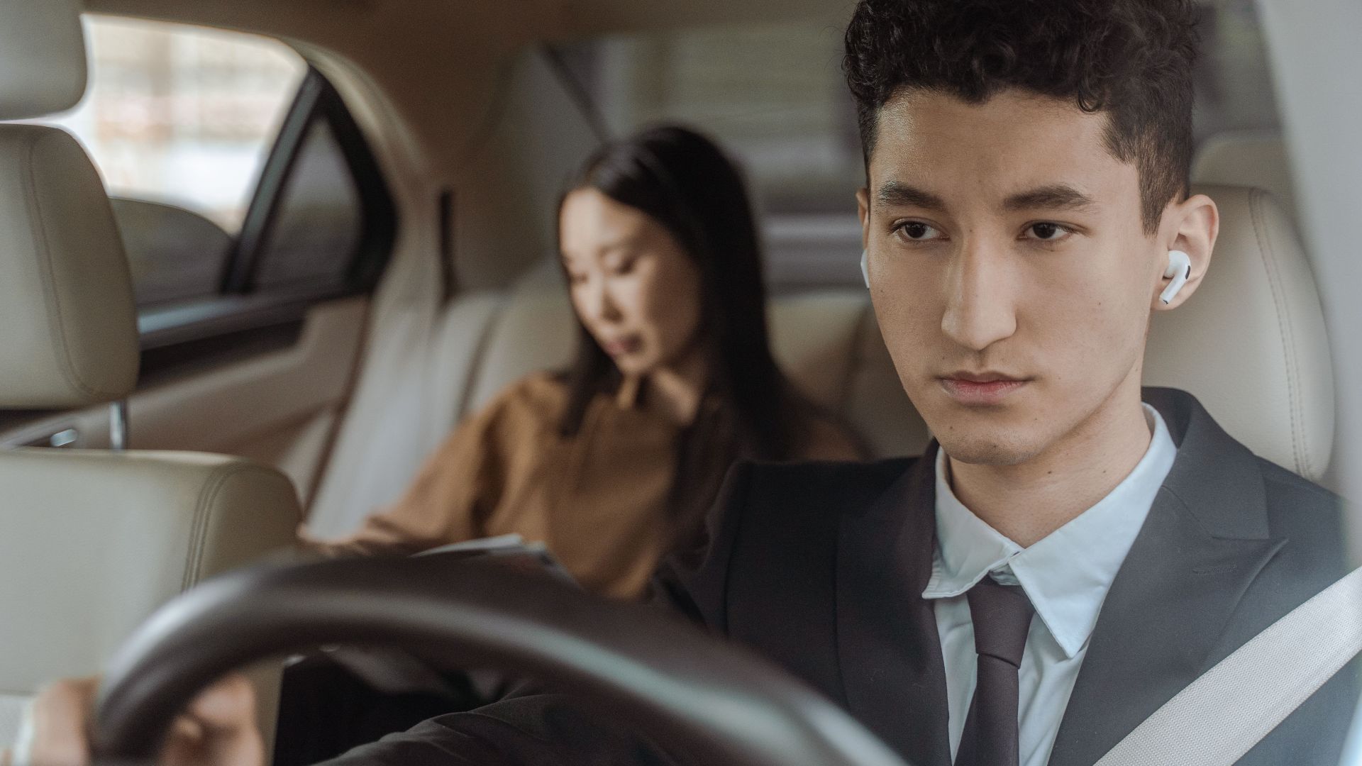 Young professional driver wearing suit and earpiece in luxury car interior with female passenger.