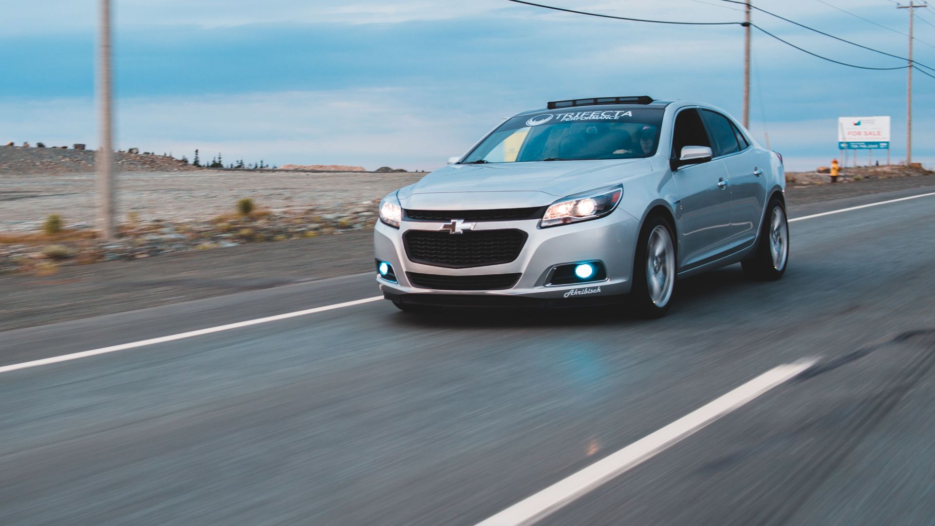 Modern car with glowing headlights and front fog lights driving on highway alongside field and utility poles