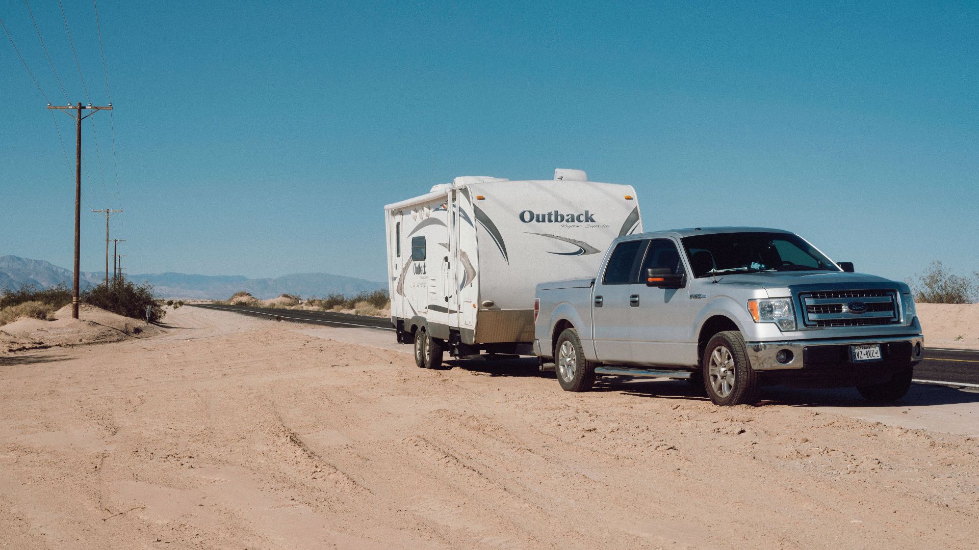 Silver truck towing an Outback trailer on a desert road under clear blue skies.