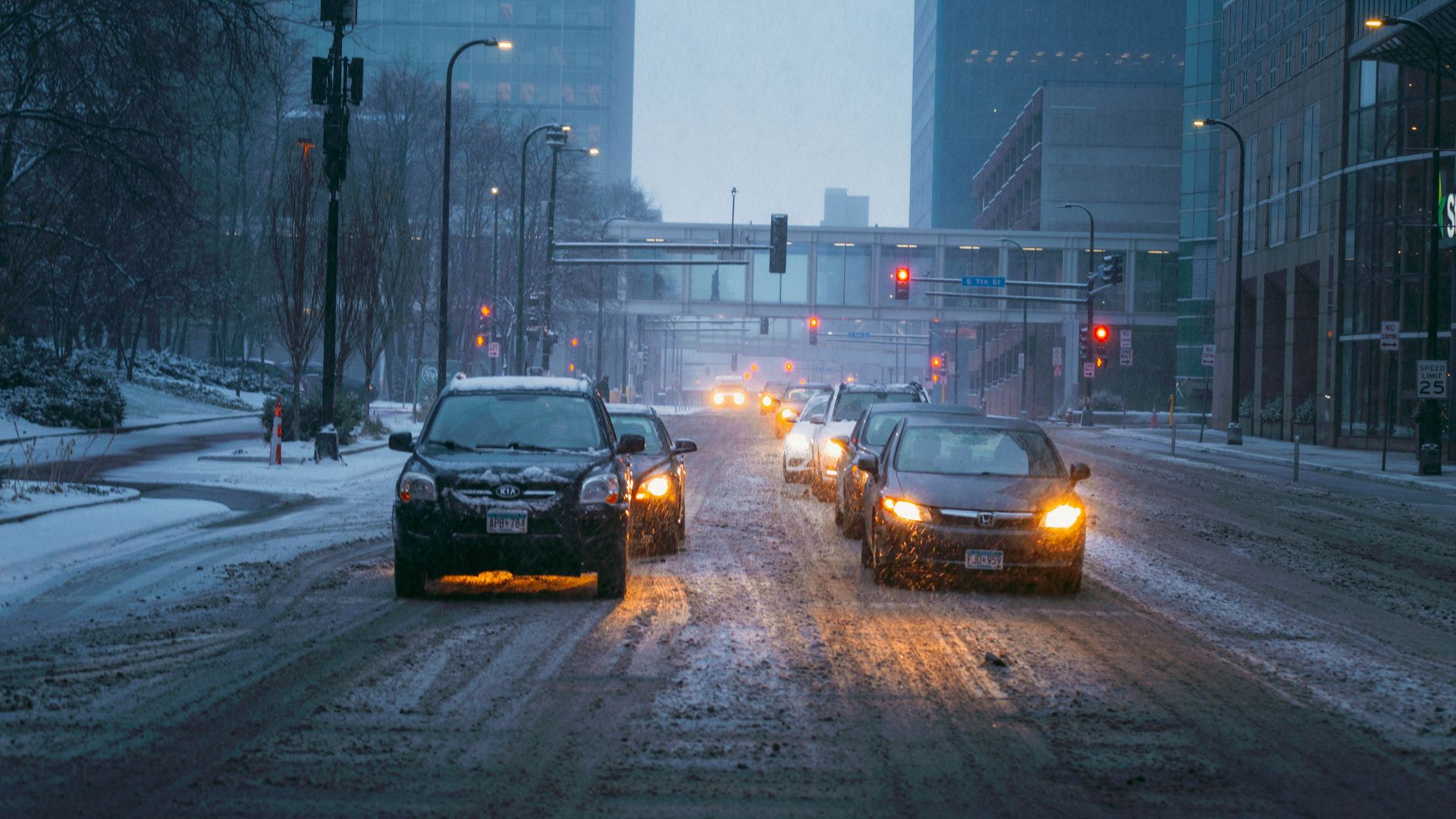 Cars driving on a snow-covered city street during an evening snowfall, with buildings lining the street.