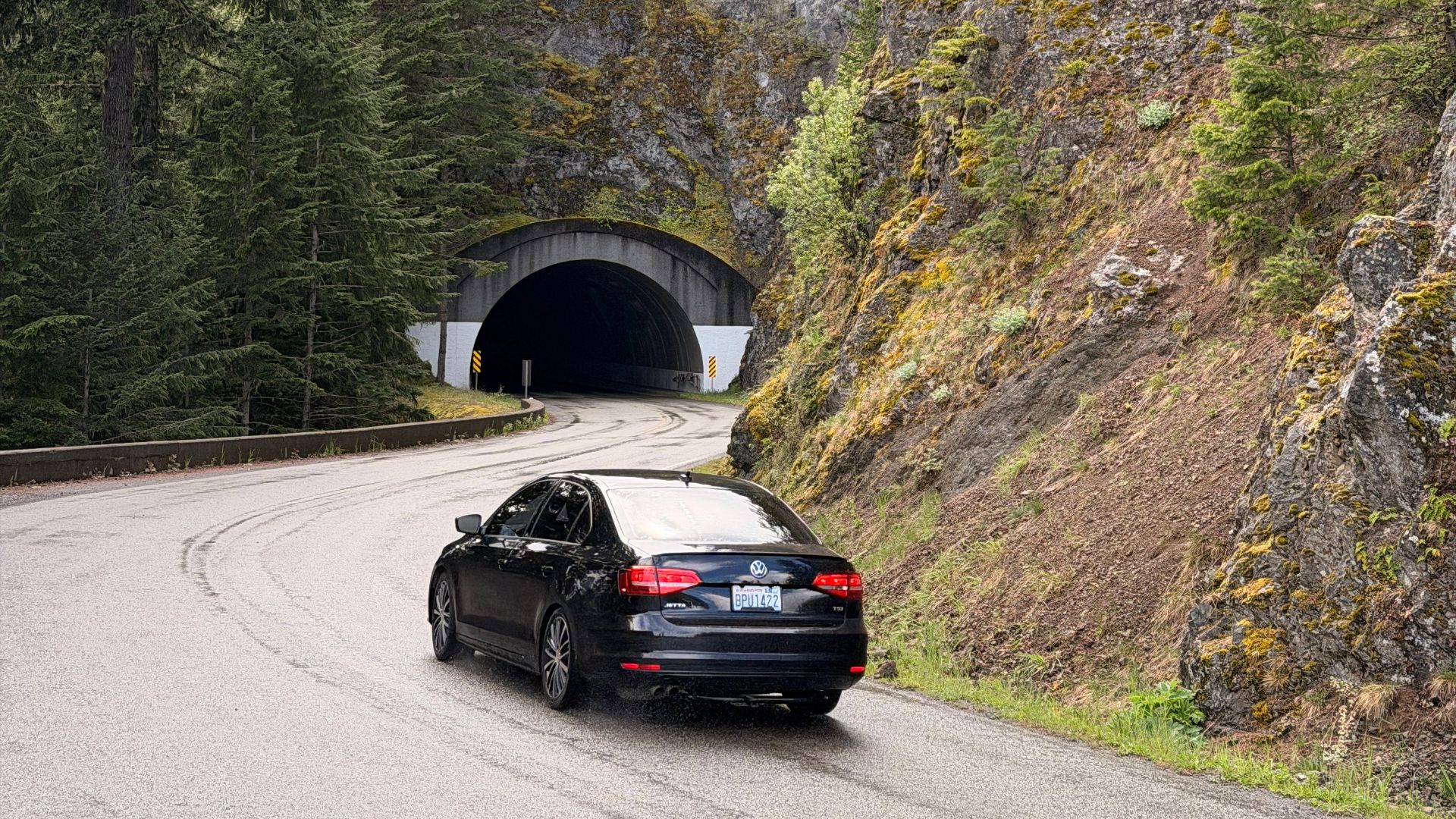 A sleek black car drives towards a mountain tunnel on a curvy road surrounded by lush greenery.
