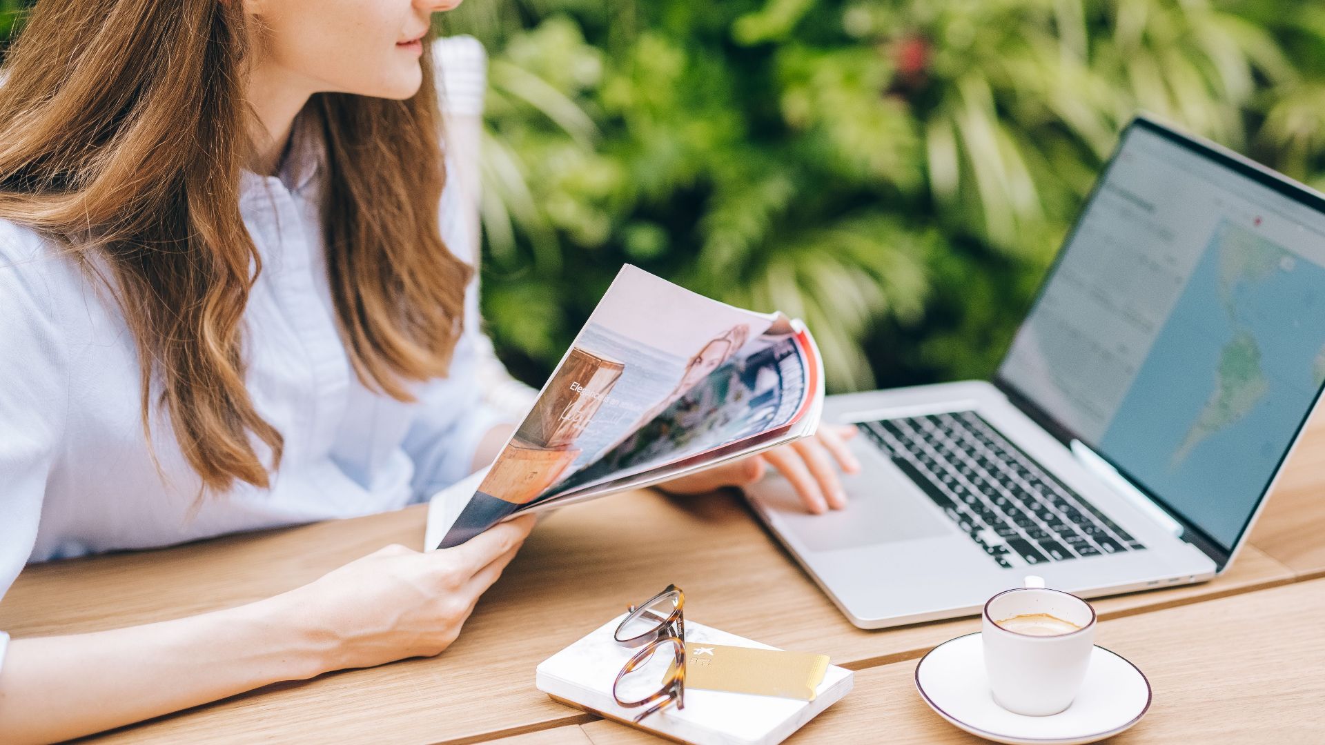 A woman sits outdoors, enjoying a magazine with coffee and laptop nearby.