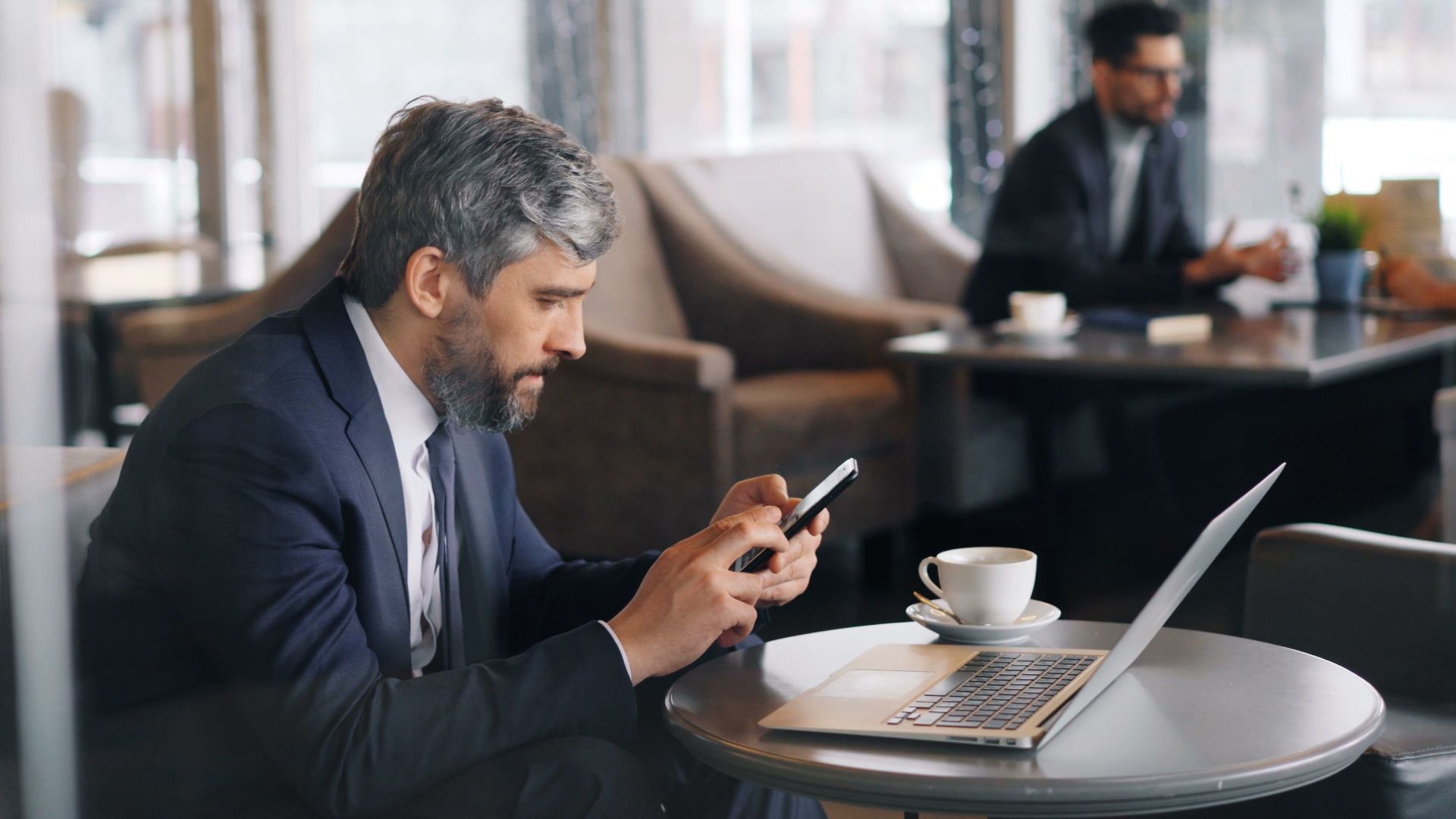 Professional man using smartphone and laptop in a café setting.