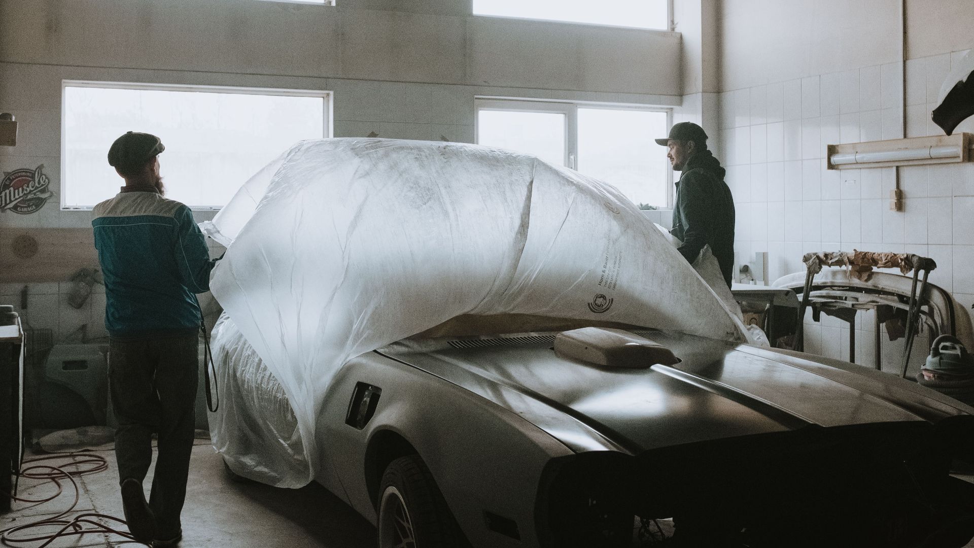 Two mechanics working on restoring a classic car in an auto garage with ample lighting.