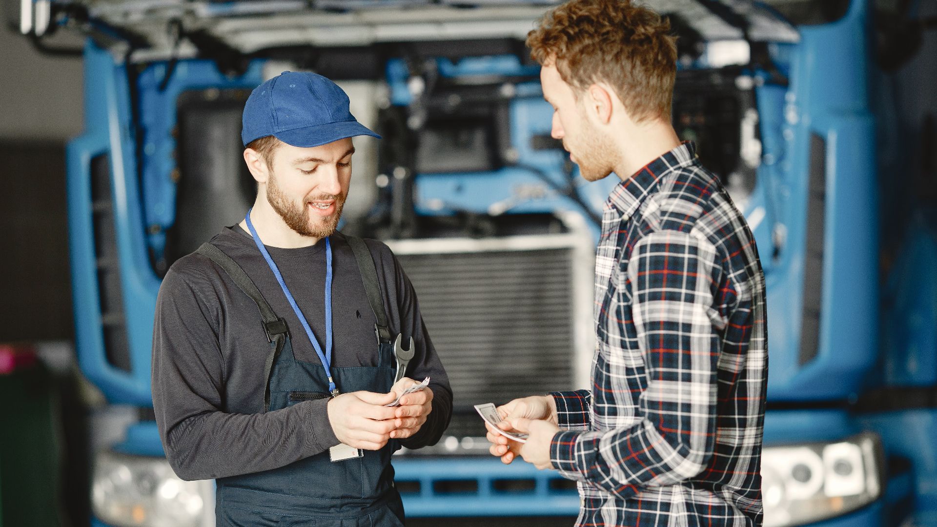 Auto mechanic in blue uniform receives payment from customer in a garage setting with a truck in the background.