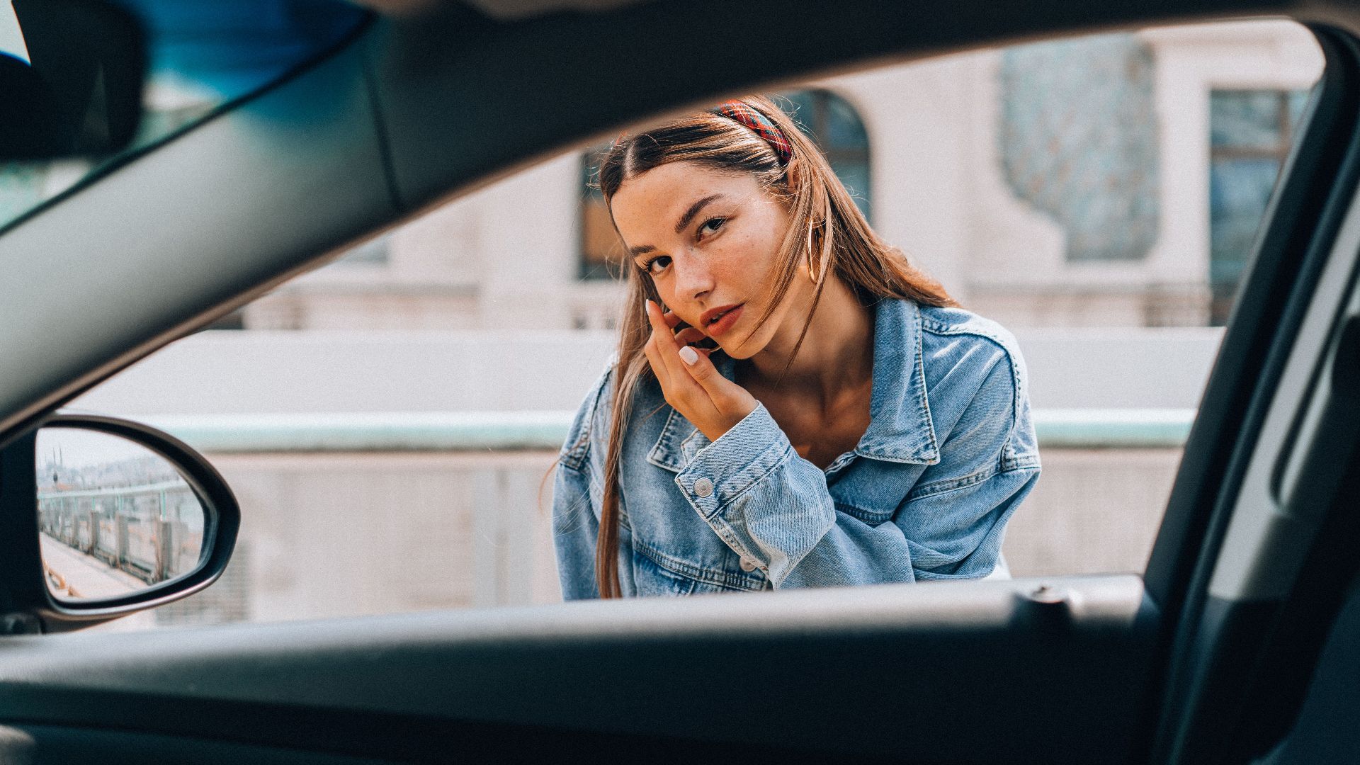 Woman in a denim jacket posing by a car window during the day.