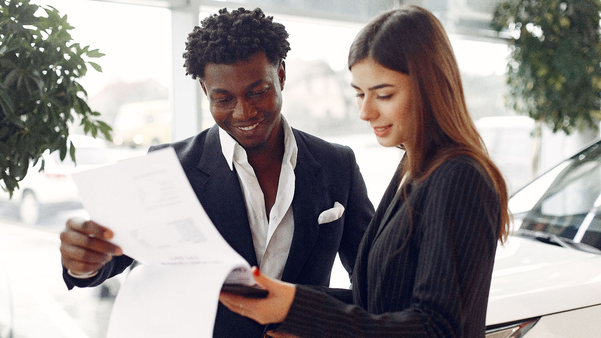 Cheerful smiling multiethnic businessman in classy suit and professional elegant female consultant standing close and reading contract details in car showroom