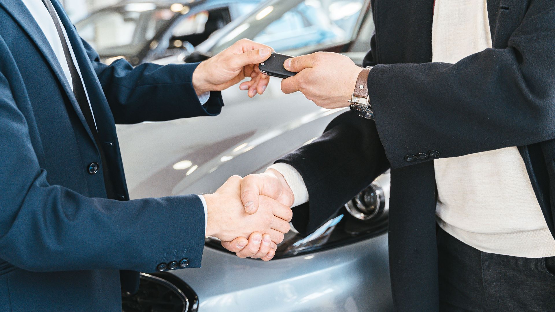 Two businessmen shaking hands and exchanging car keys in a dealership. Symbolizes a successful deal.
