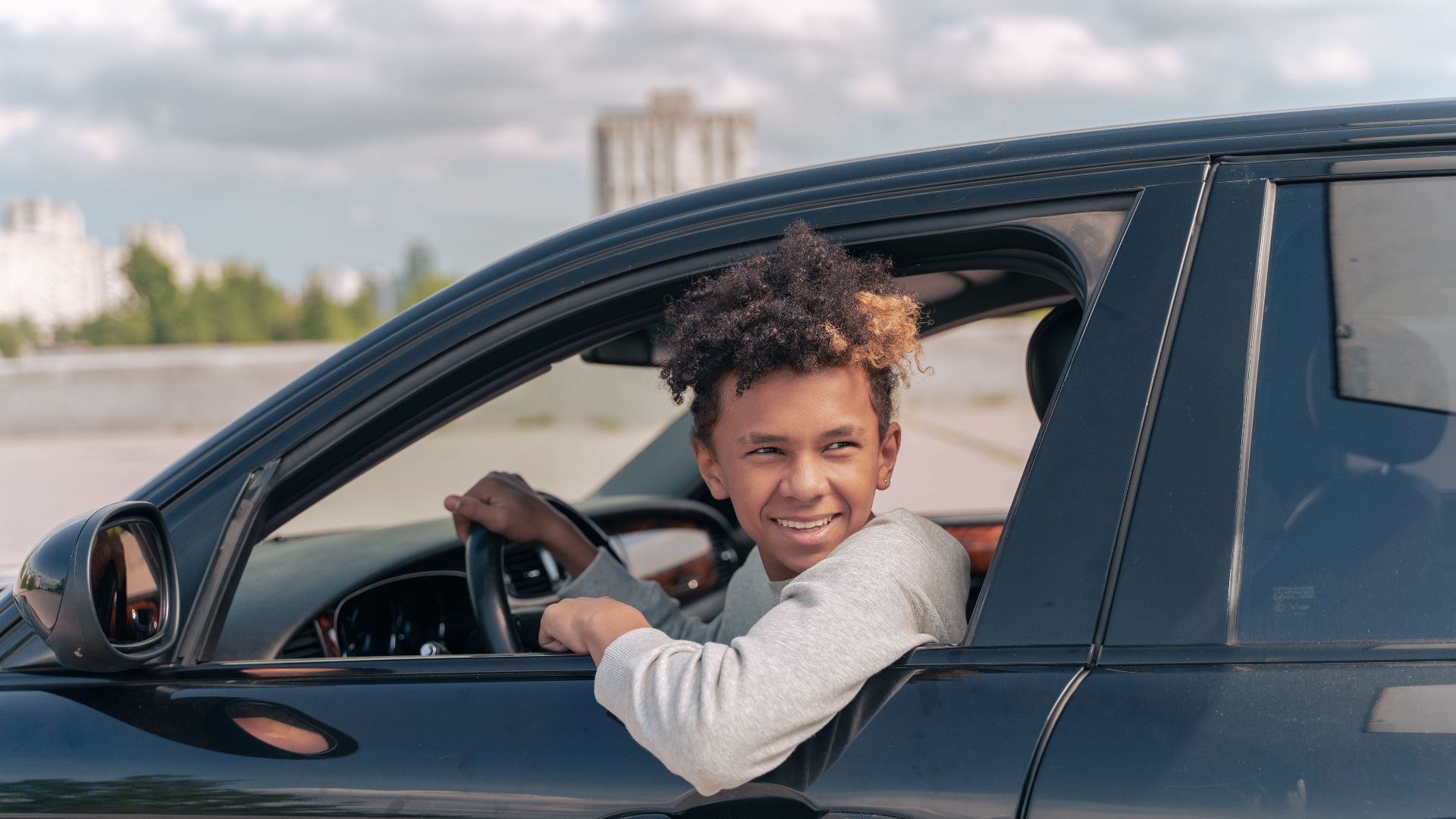 Teenager with curly hair smiling while sitting in car outdoors on a sunny day.