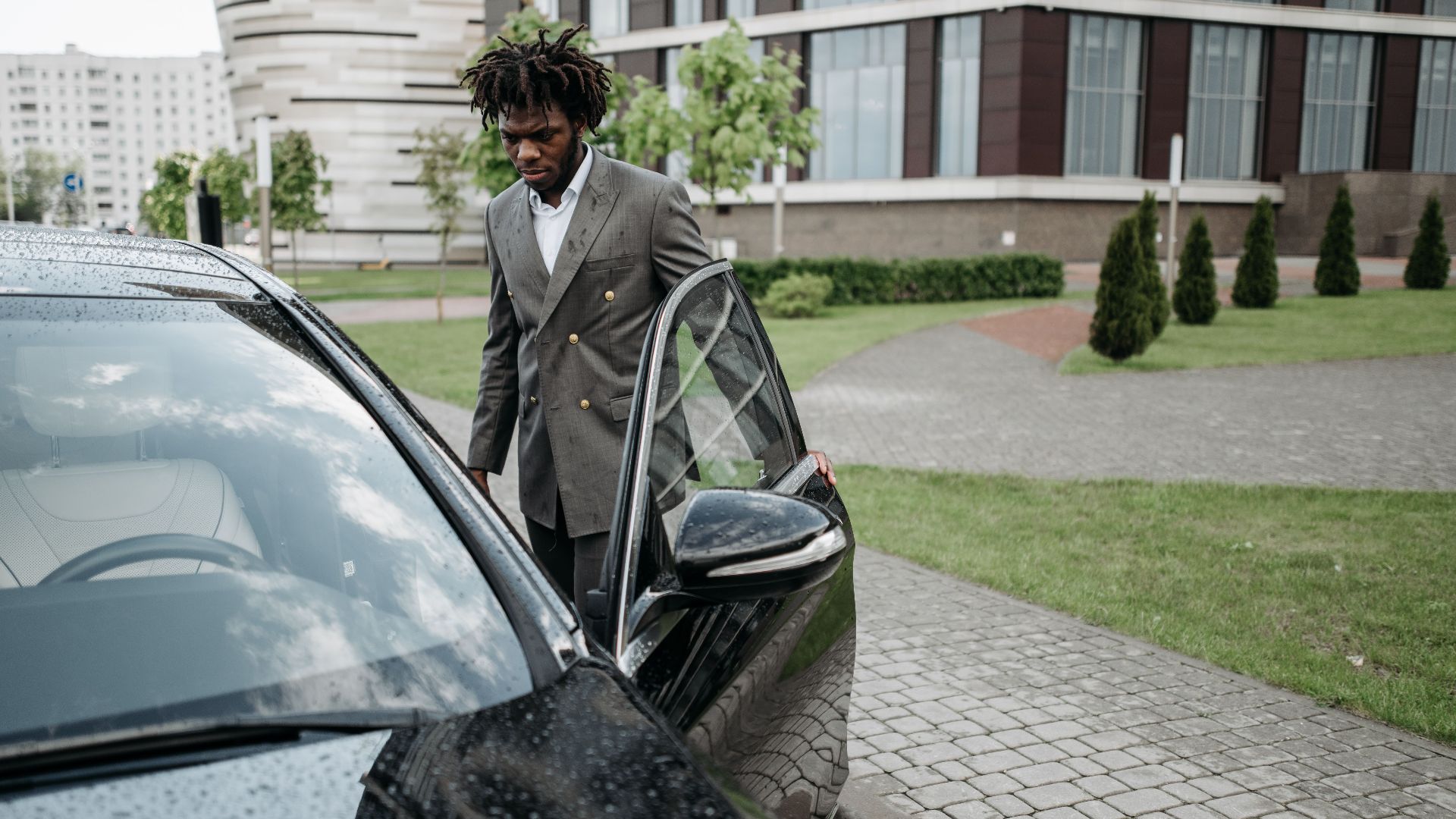 Man in a gray suit opening a black car door in an urban setting.