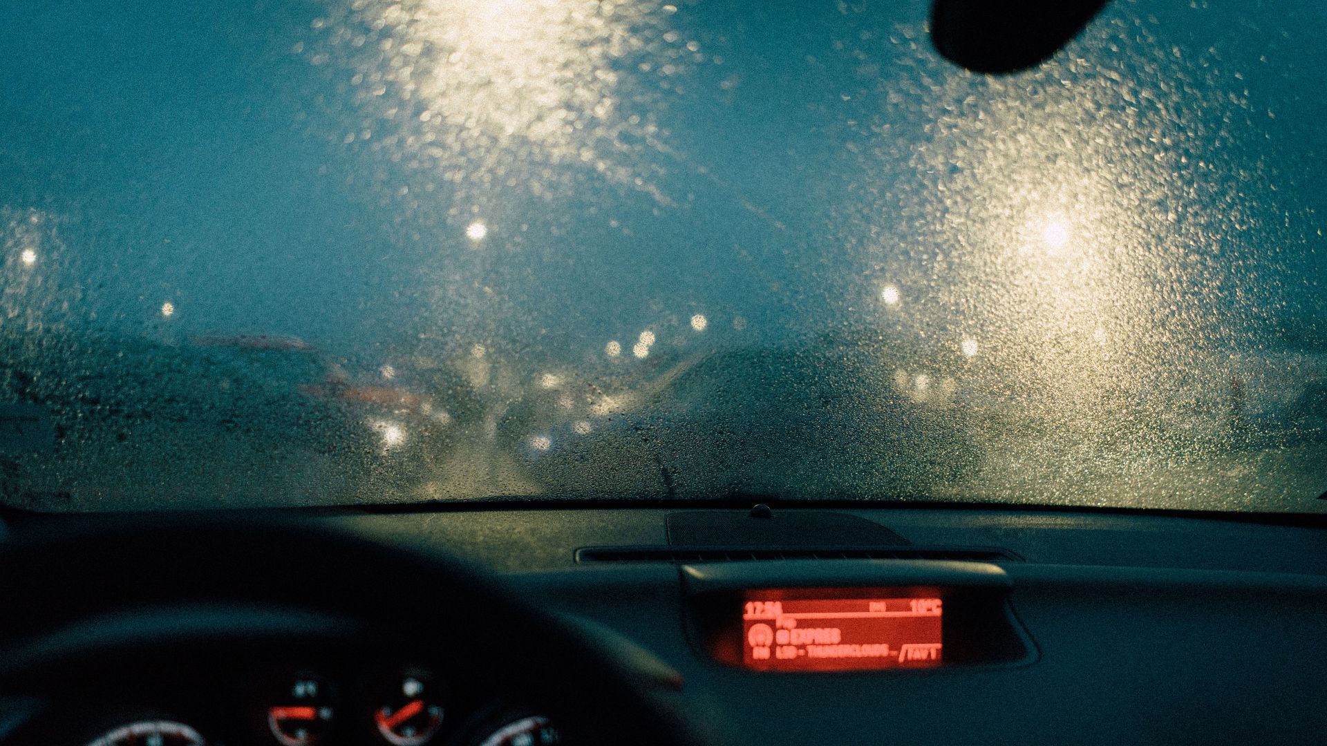View from car interior with raindrops on windshield and blurred lights at night.