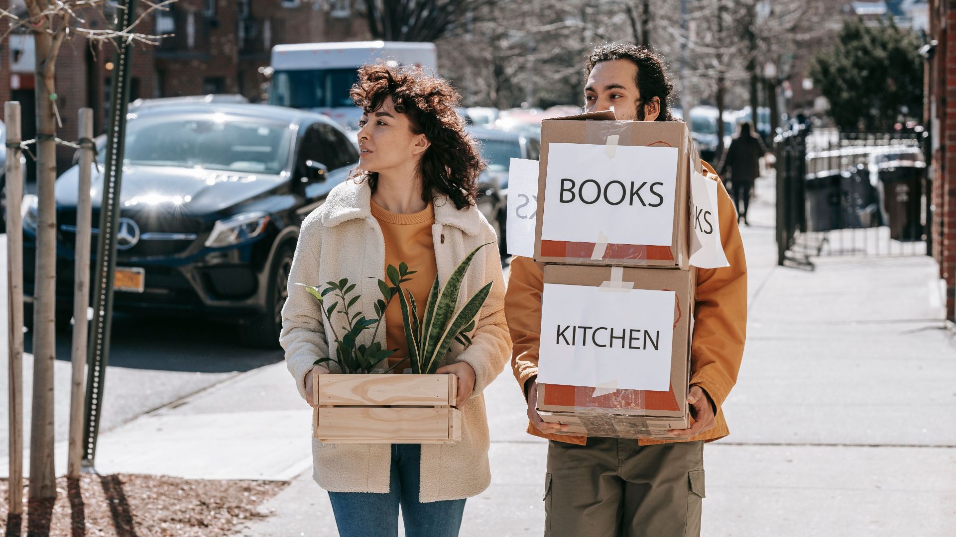 A couple walks in a city street carrying boxes labeled 'Books' and 'Kitchen' symbolizing relocation.