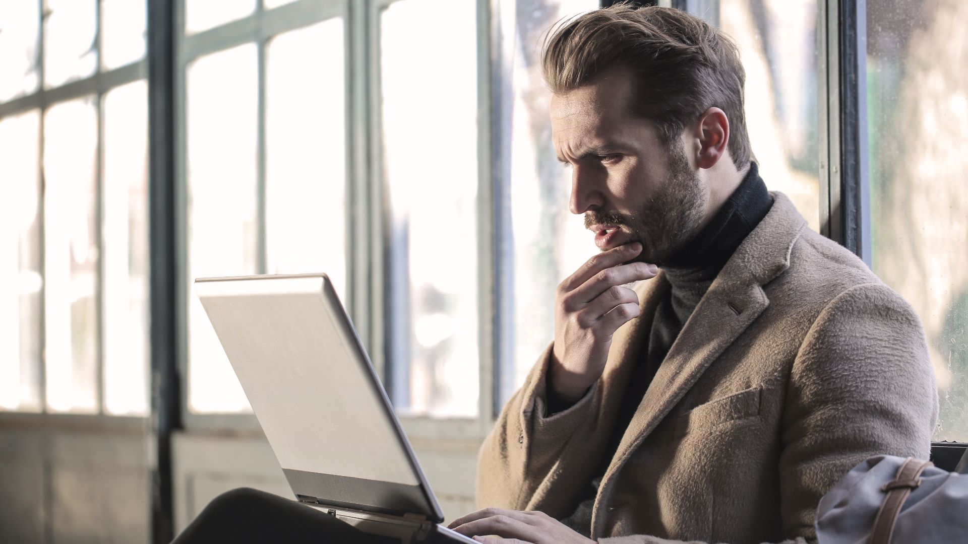Man looking contemplative while working on a laptop in a well-lit indoor space.