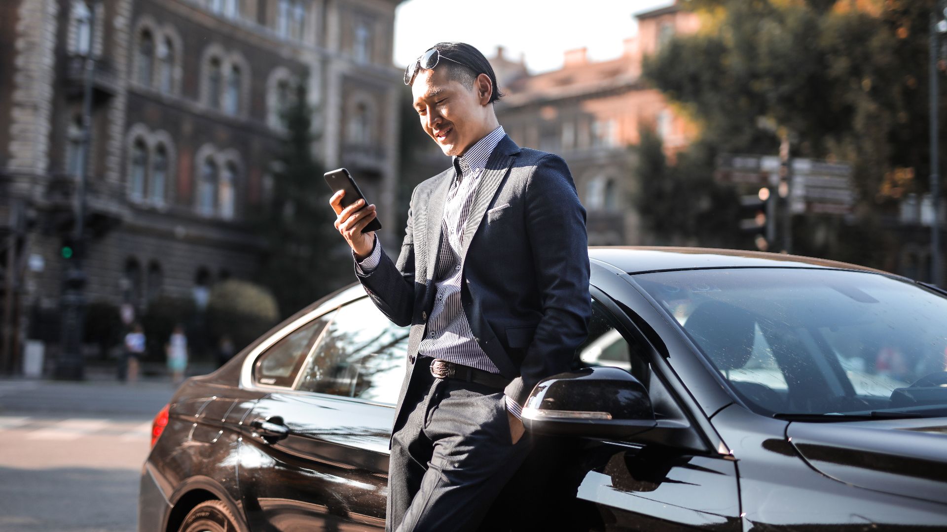 Smiling businessman in a black suit texting on his phone while leaning against a parked car in the city.