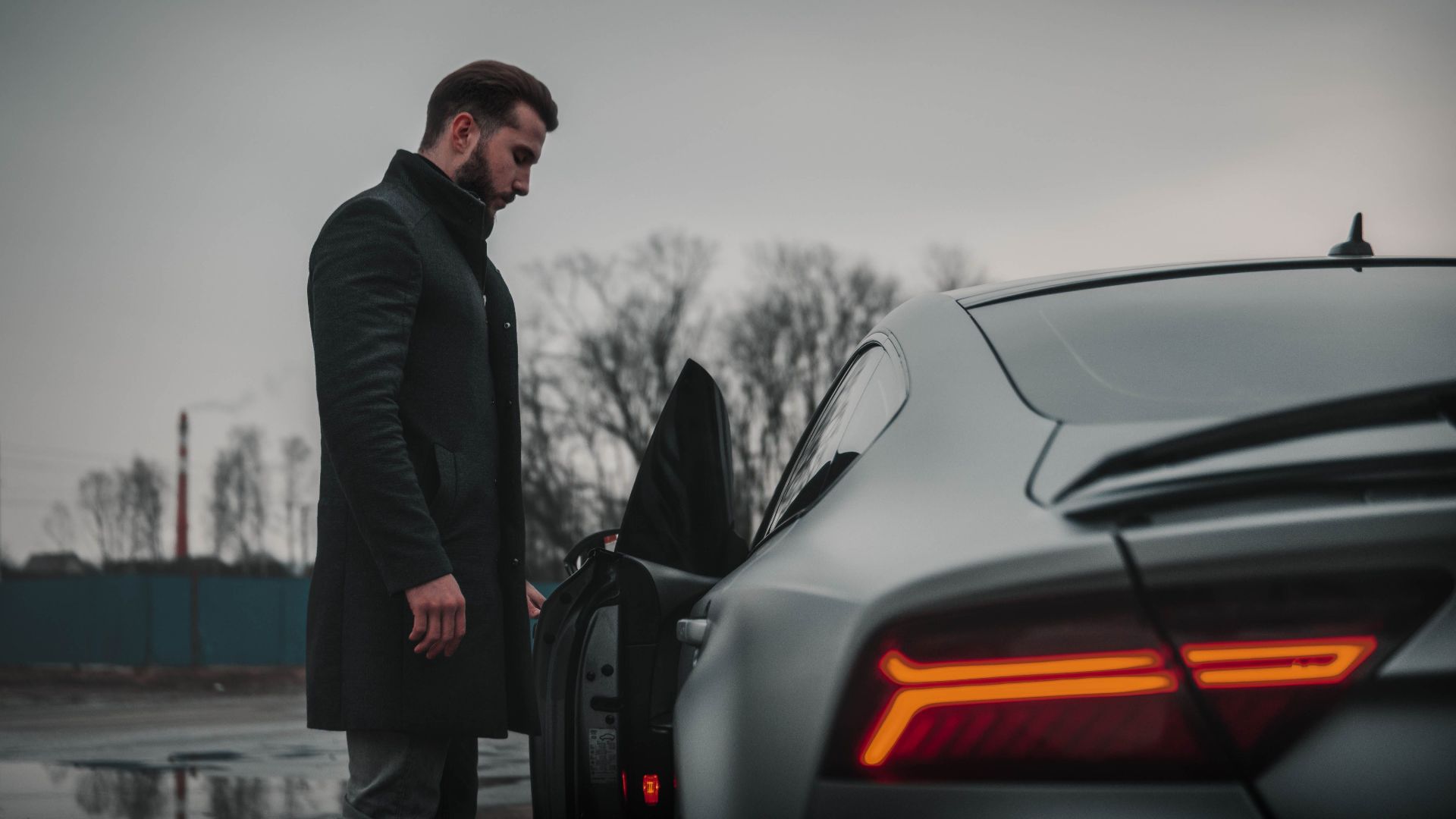 A man in a black coat stands beside a luxury car on a cloudy day in Moscow, Russia.