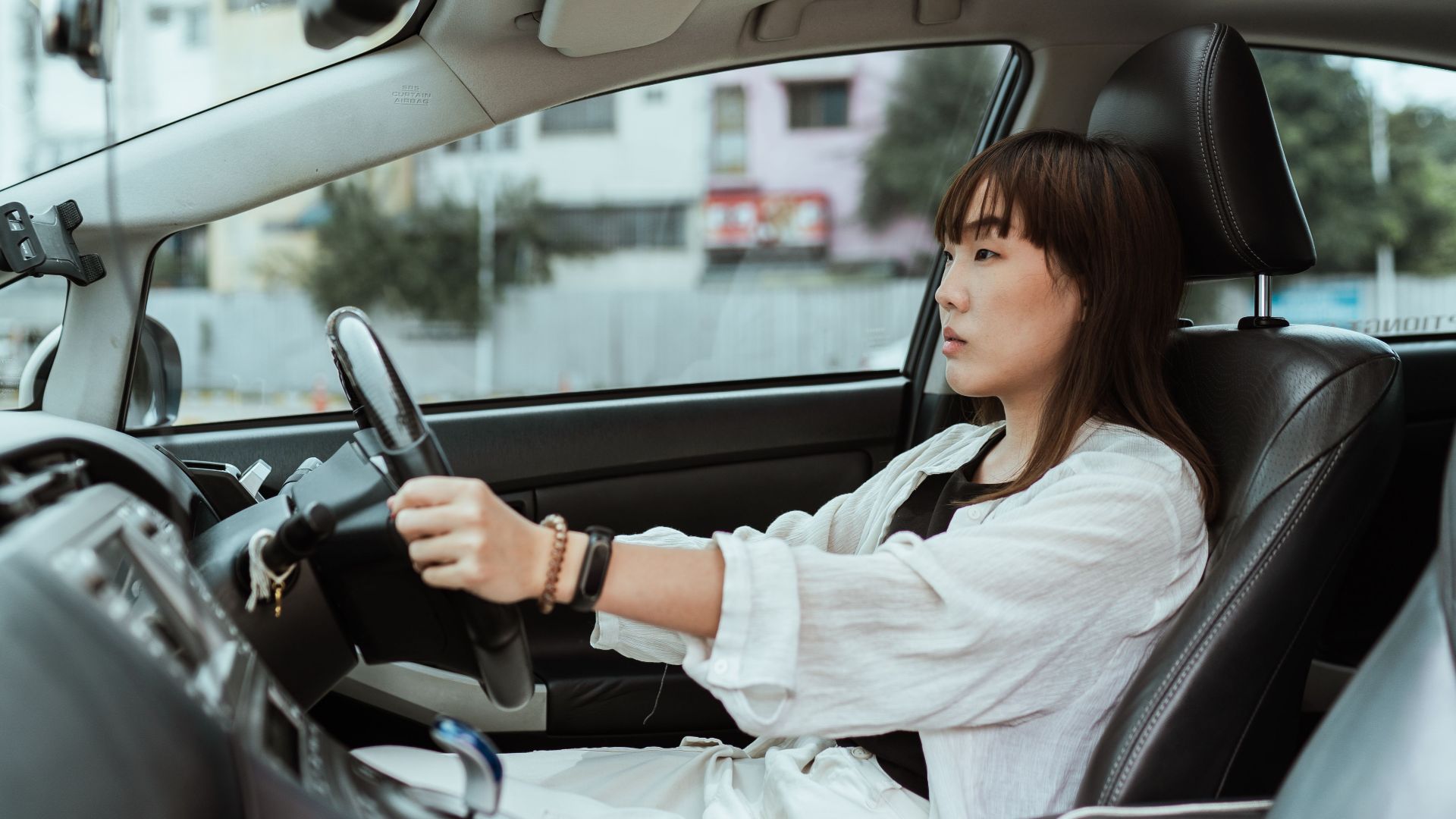 Side view unemotional Asian female in casual white clothes steering right hand drive car along urban streets in daytime