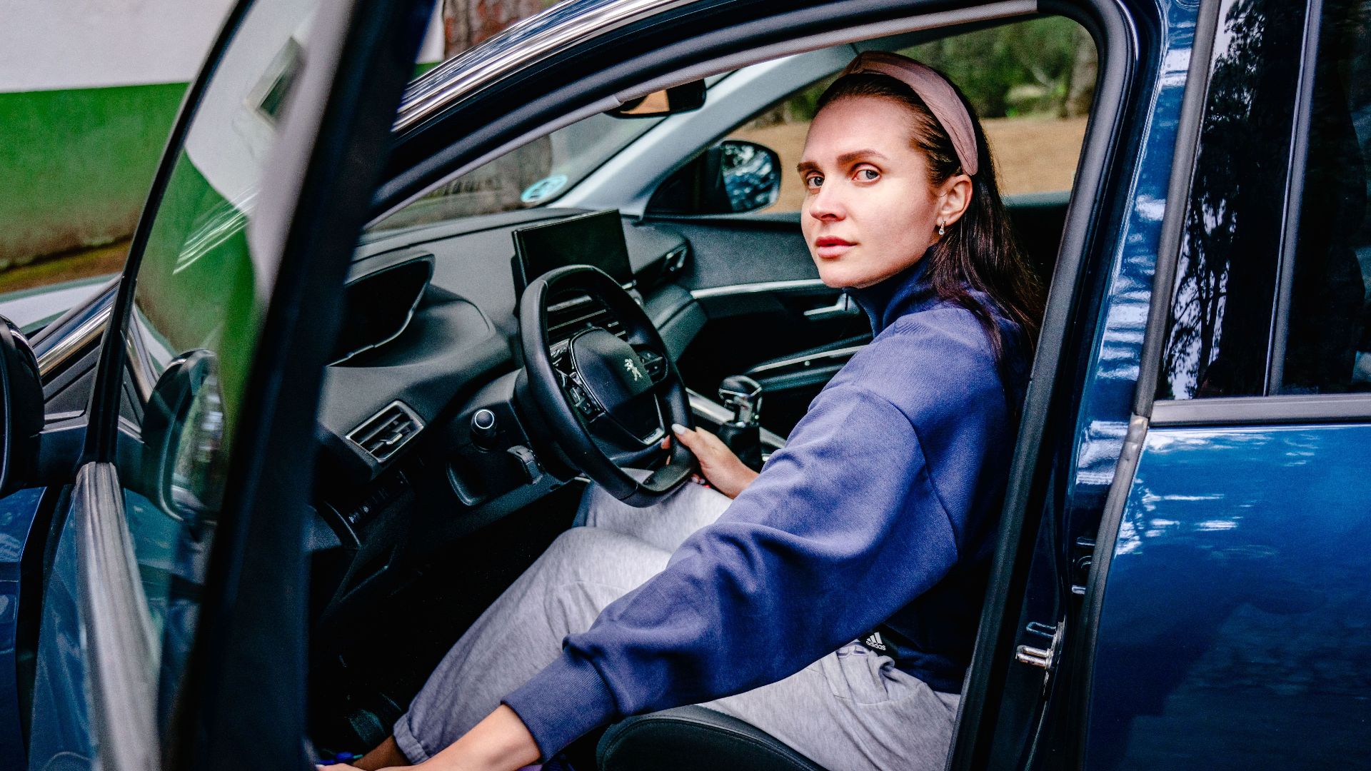 Woman sitting in the driver's seat of a car with an open door, looking outside. Natural setting.