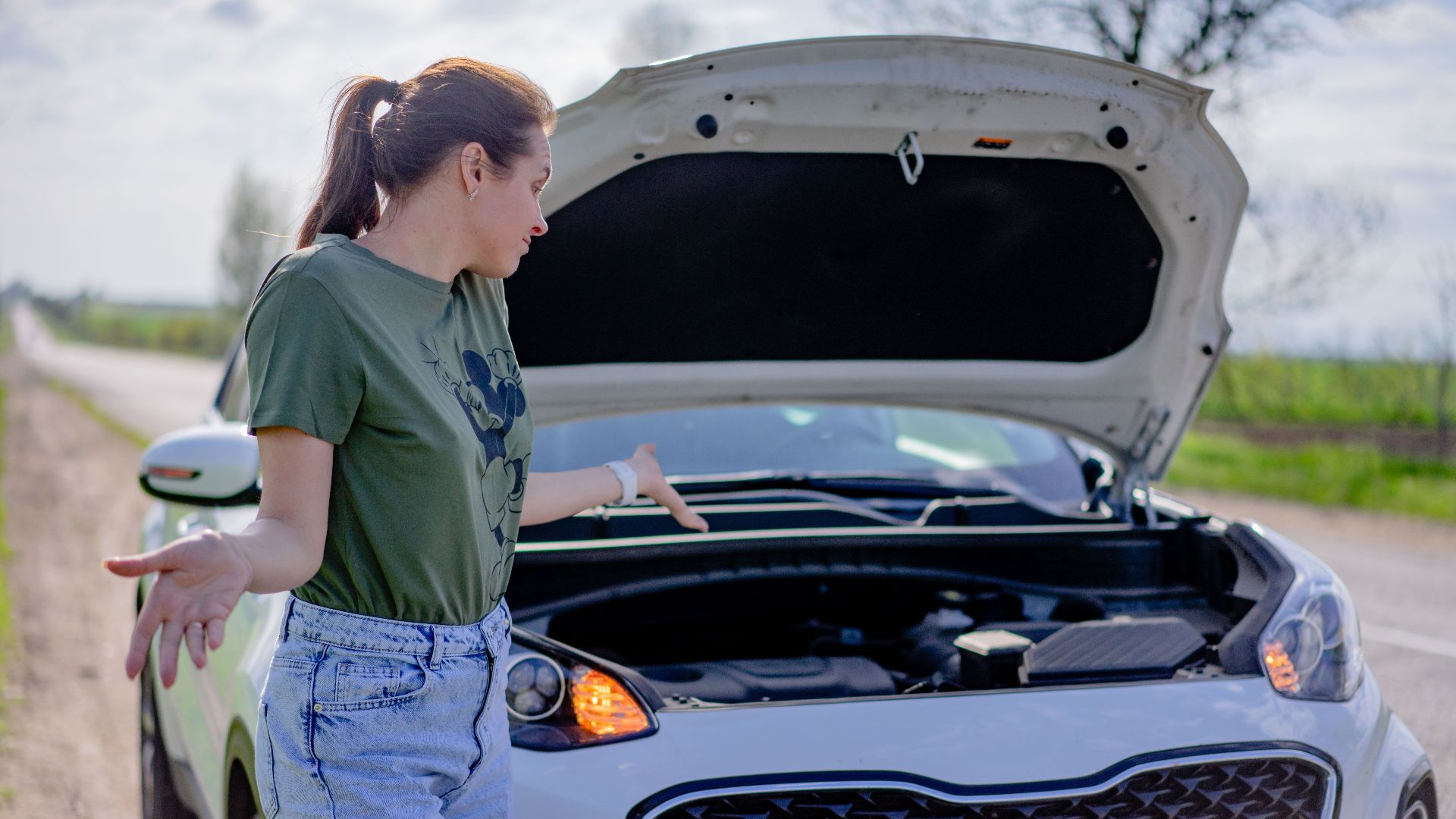 A woman looks puzzled by her broken car with an open hood on a rural road.