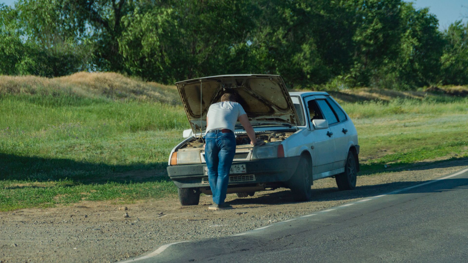 A man stands by his car on the roadside, inspecting a breakdown issue.